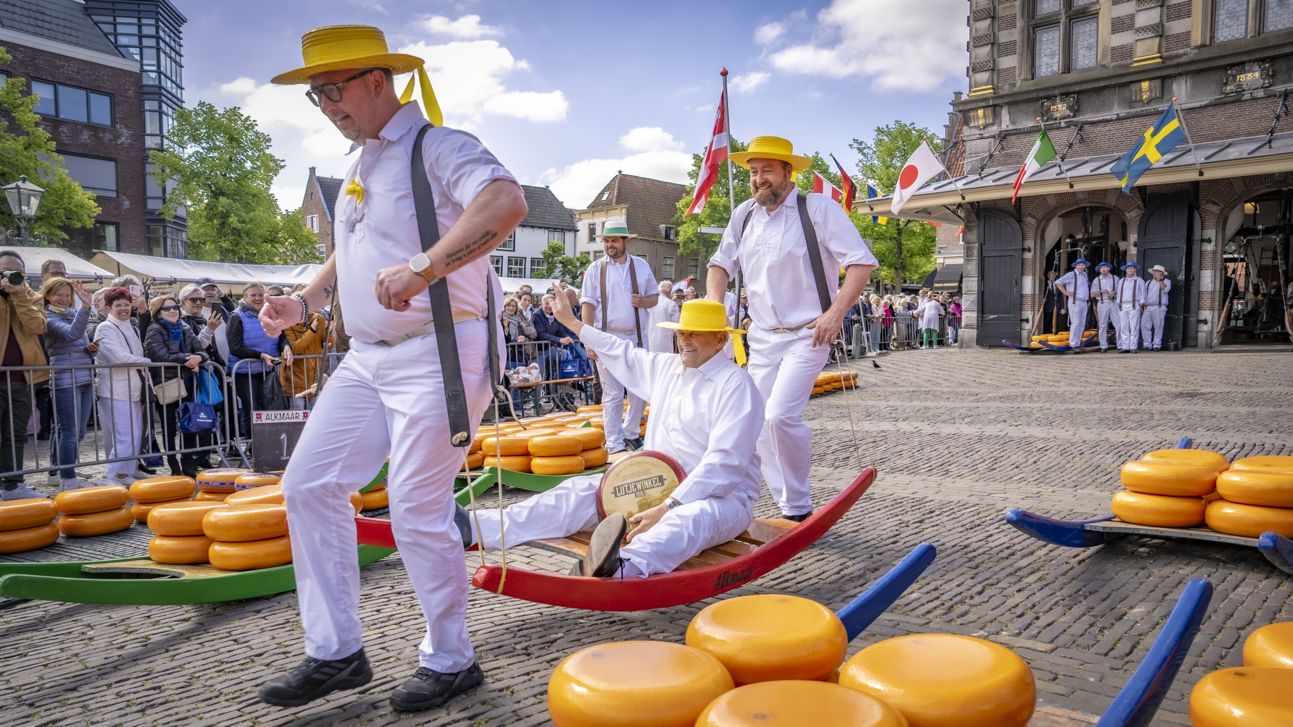 Mannen in traditionele witte kleding en gele hoeden verplaatsen kaasen op een drager op de kaasmarkt in Alkmaar, omgeven door toeschouwers.
