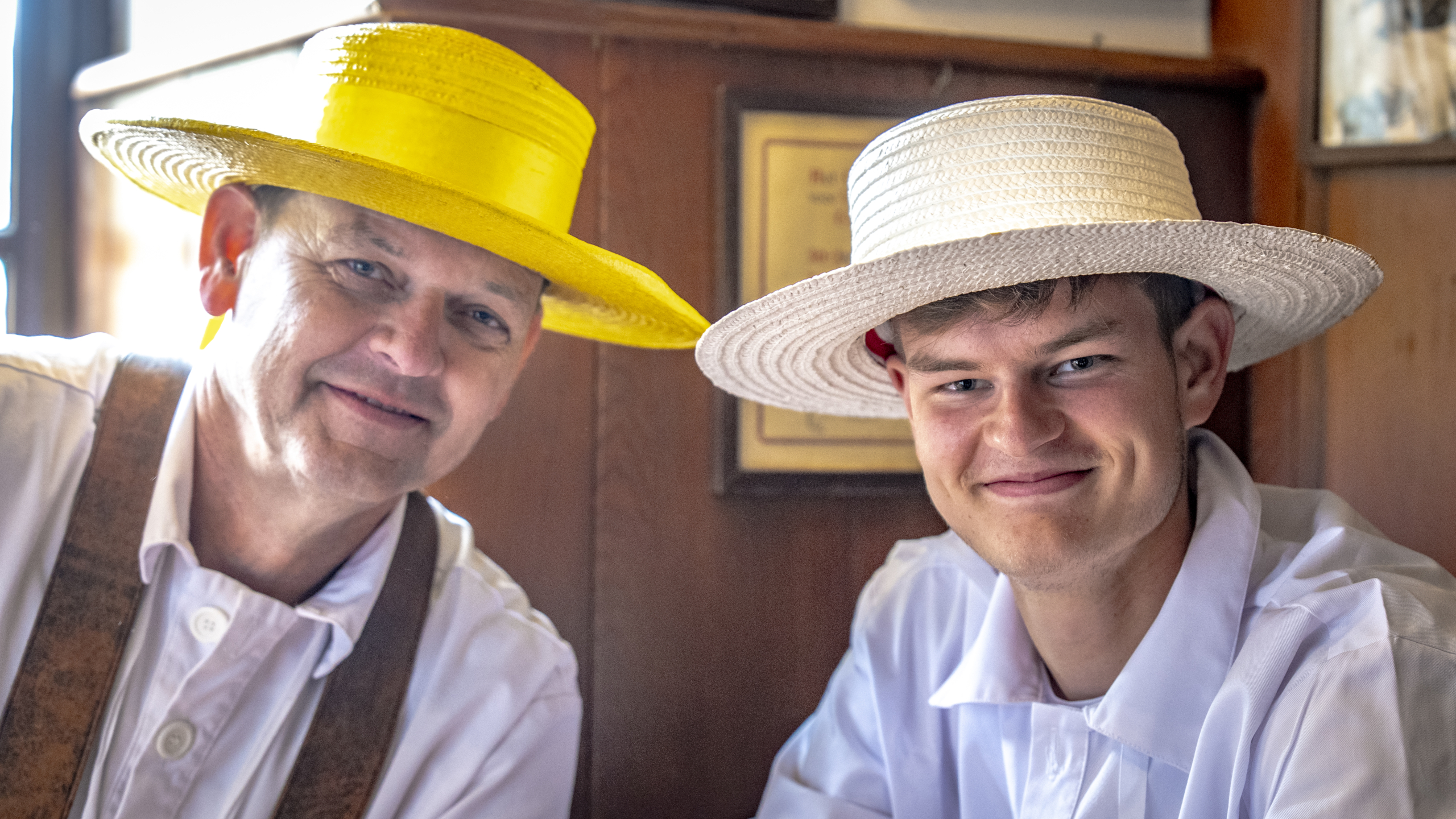 Twee mannen dragen witte tops en strohoeden, de linker is geel en de rechter wit, terwijl ze naar de camera glimlachen in een houten interieur.