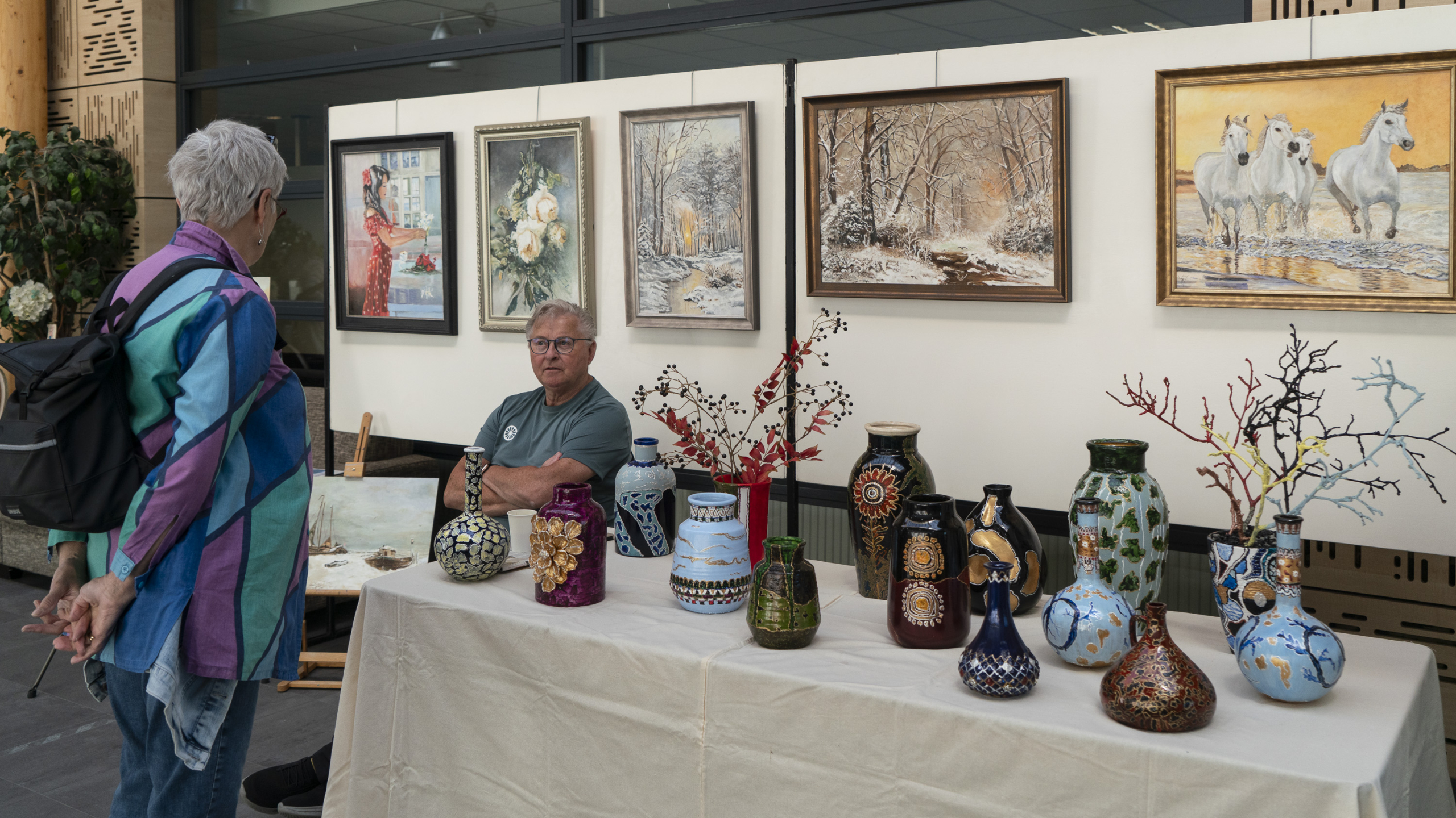 Een man zit aan een tafel vol met versierde vazen terwijl hij een vrouw aanspreekt bij een kunsttentoonstelling, met schilderijen op de achtergrond.
