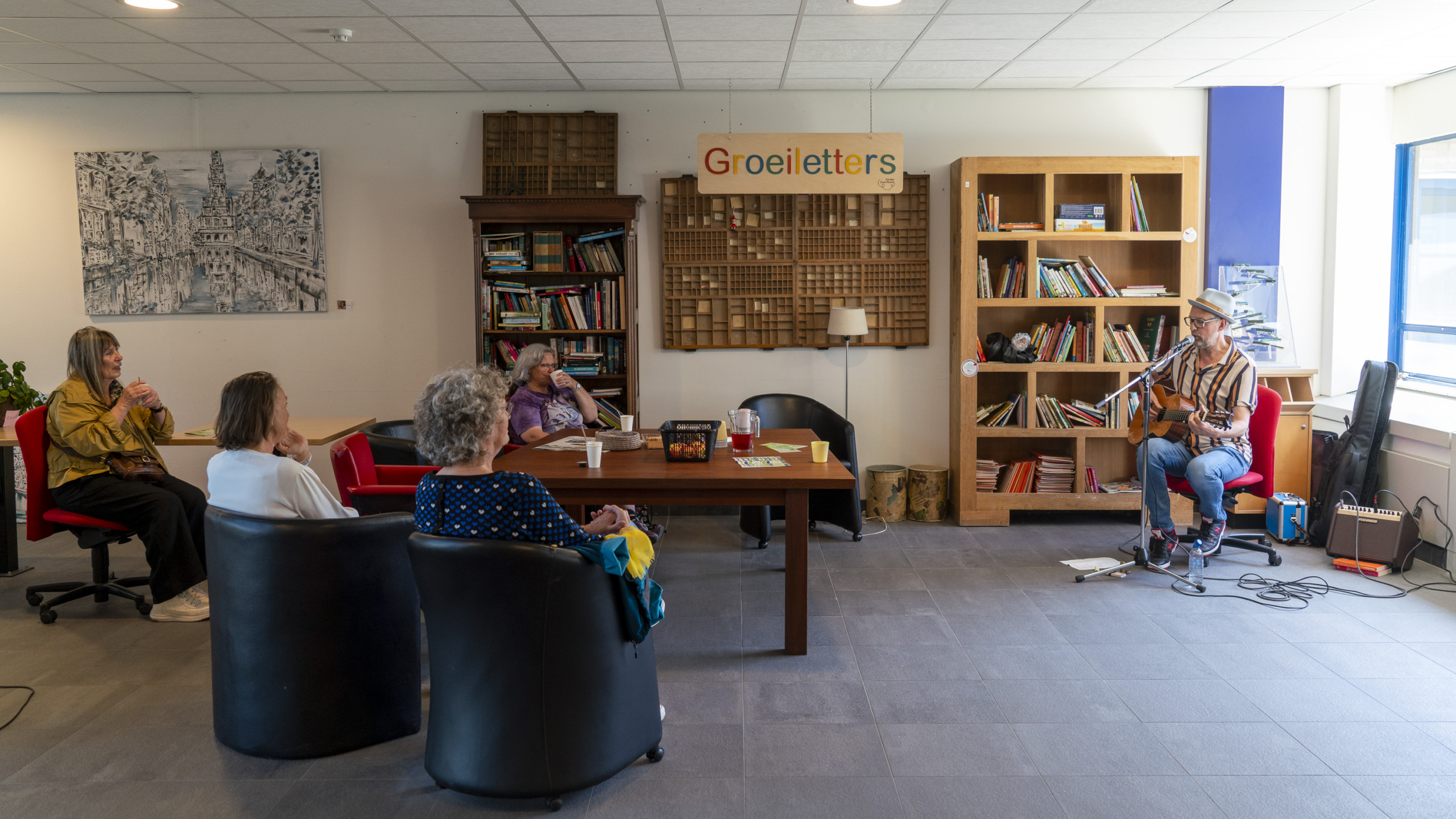 Een muzikant speelt gitaar voor een kleine groep mensen in een kamer met boekenplanken en een schilderij aan de muur met de tekst 'Groeiletters'. Er is een tafel met bekers en een kan, en de luisteraars zitten op stoelen rondom de tafel.