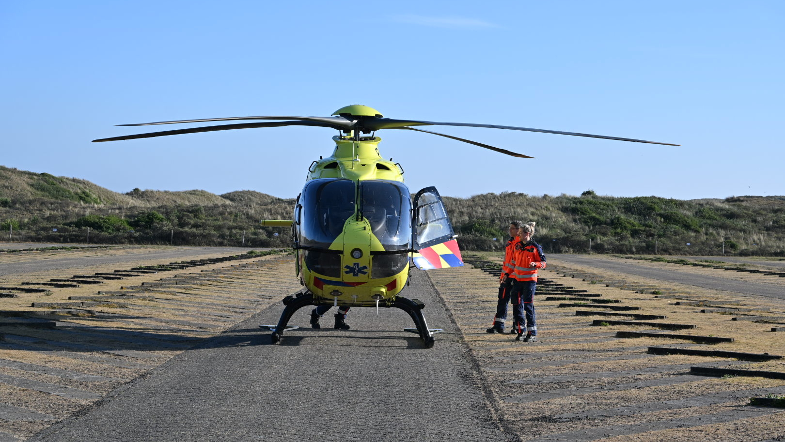 Een gele medische traumahelikopter staat geparkeerd op een open terrein. Twee mensen in oranje veiligheidskleding staan naast de helikopter. De achtergrond toont een heuvelachtig gebied met gras.