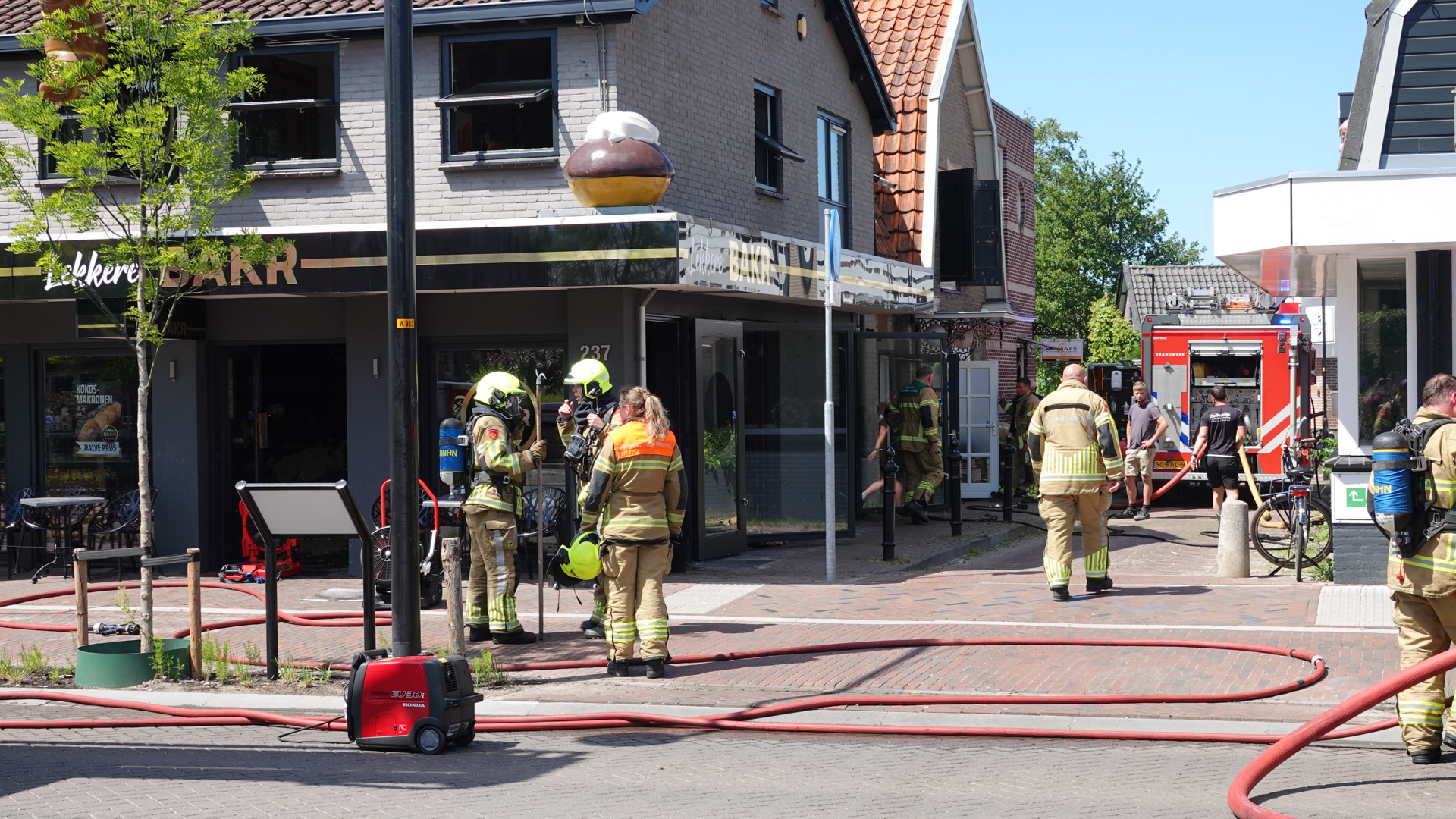Brandweerlieden in uniform staan buiten een gebouw met gebroken ramen, terwijl een brandweerwagen en slangen zichtbaar zijn op straat.