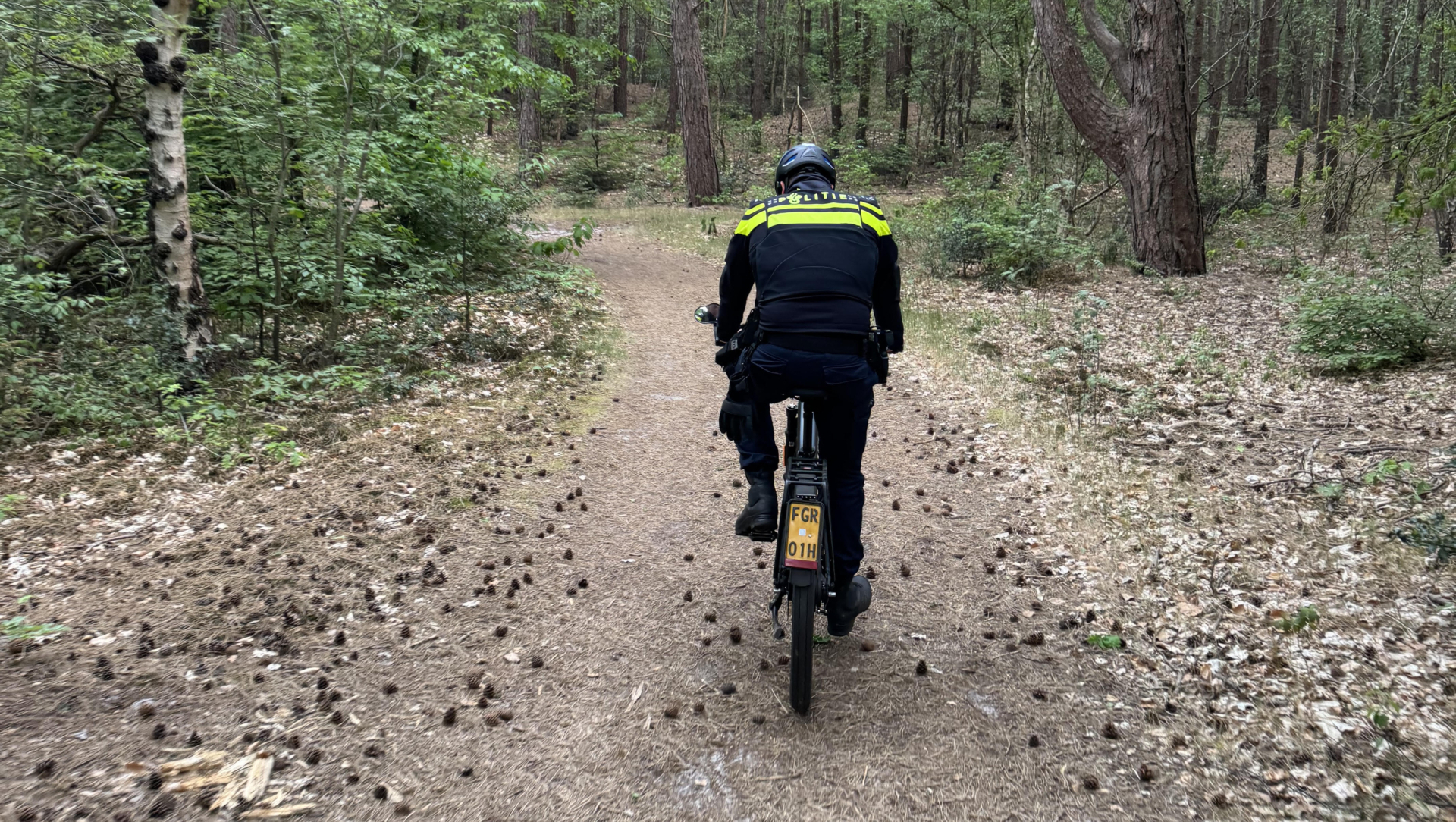 Een persoon in een donkere uniform fietst over een bospad omgeven door hoge bomen en groen.