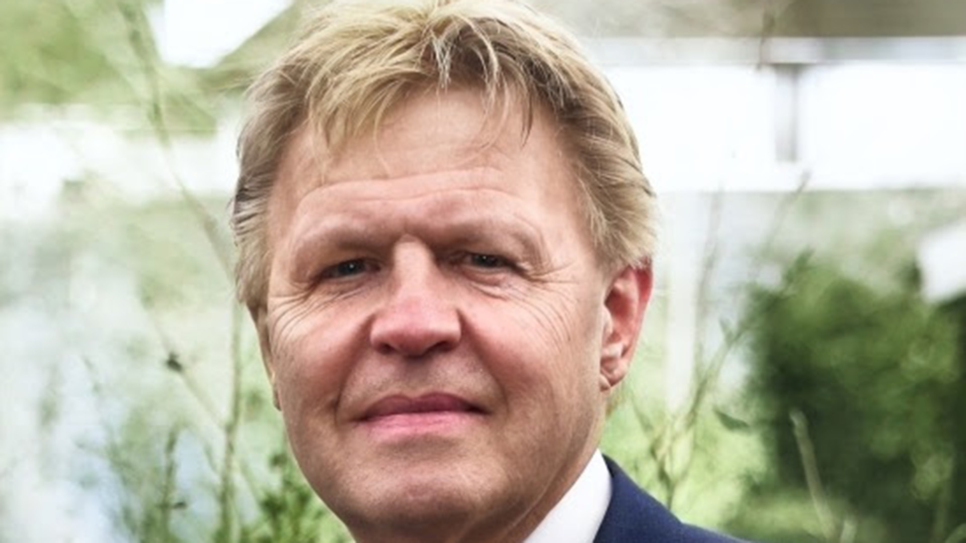 Portret van een man met blond haar en een blauwe blazer, staande voor een groene, onscherpe achtergrond.