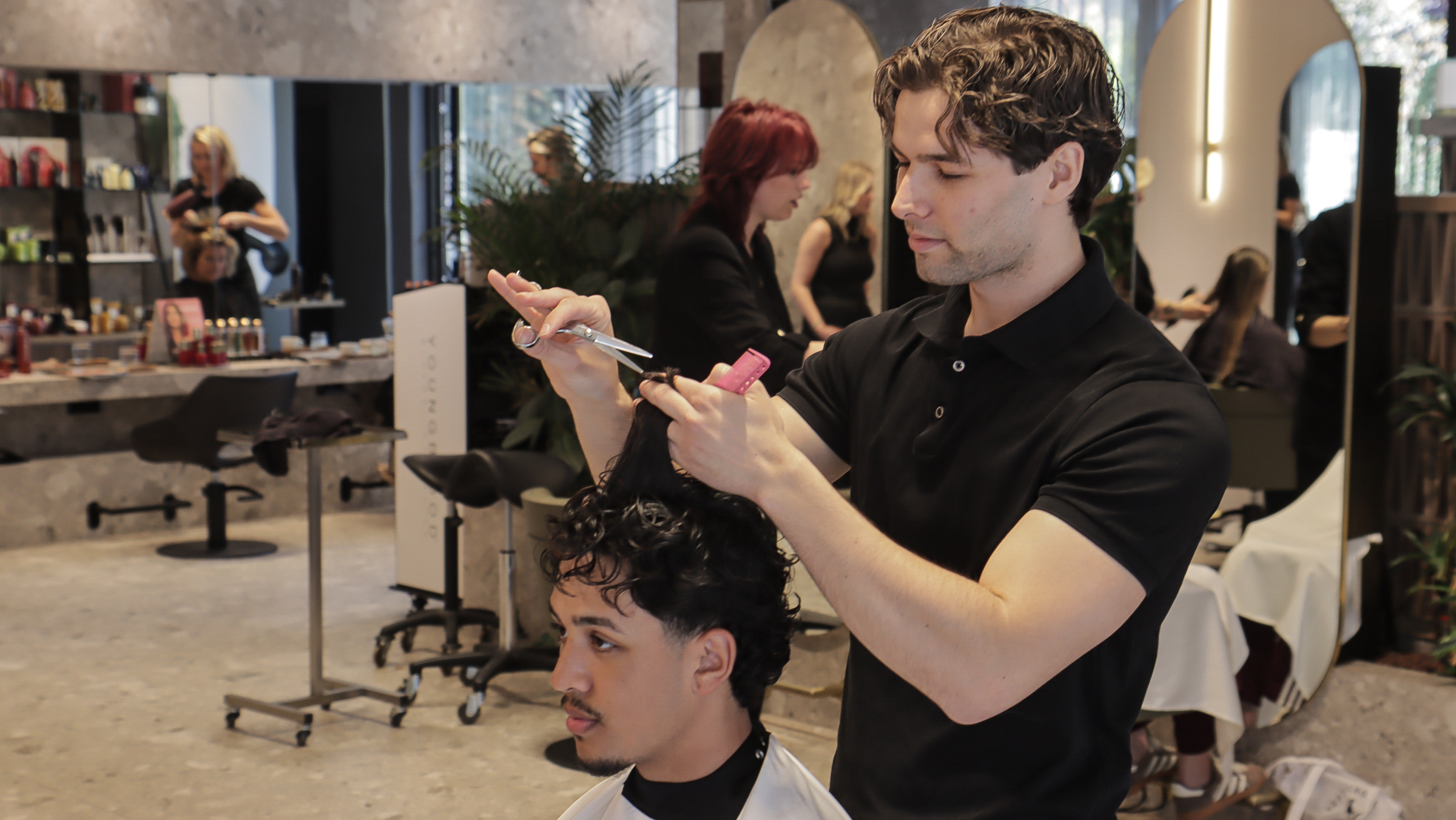 Een kapper knipt het haar van een man in een moderne kapsalon, met spiegels en diverse haarproducten op de achtergrond.