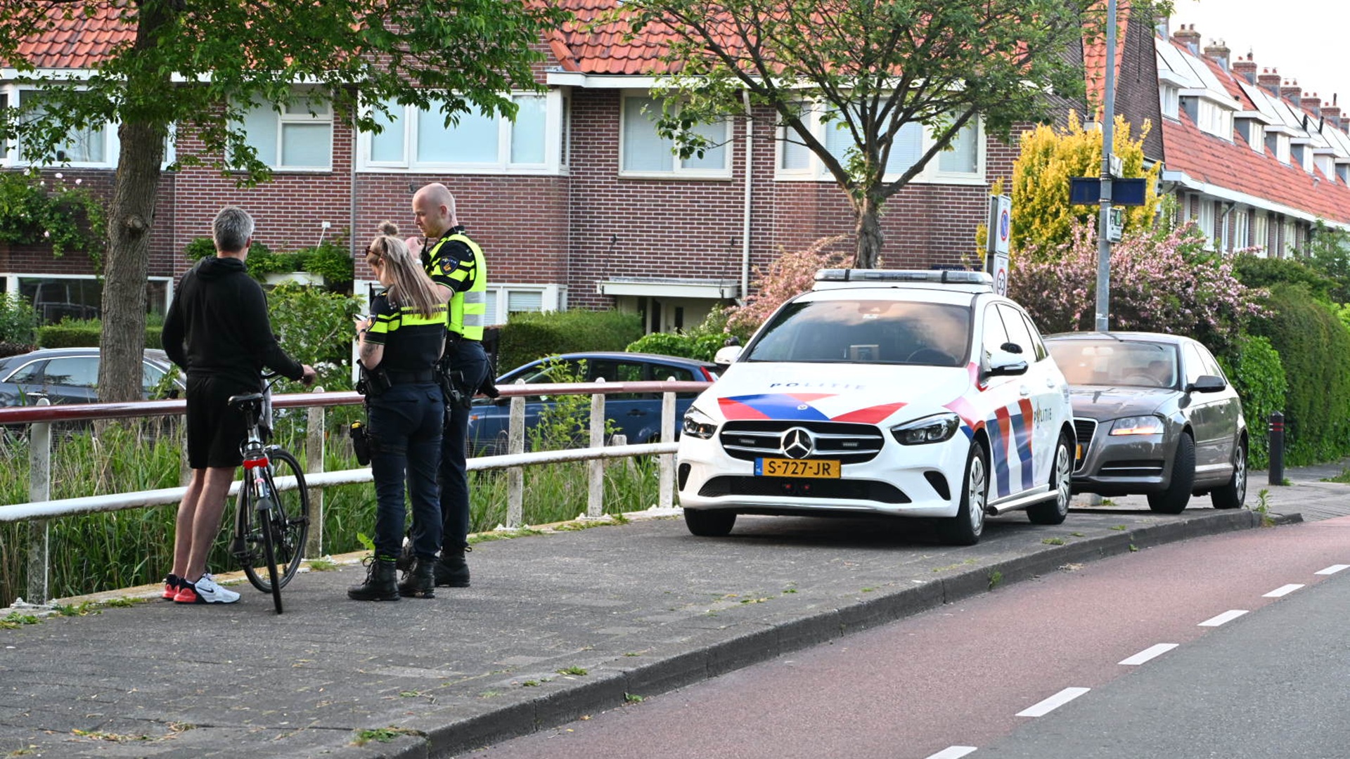 Twee politieagenten praten met een man die naast zijn fiets staat op een trottoir, naast een geparkeerde politieauto in een woonwijk met bakstenen huizen op de achtergrond.