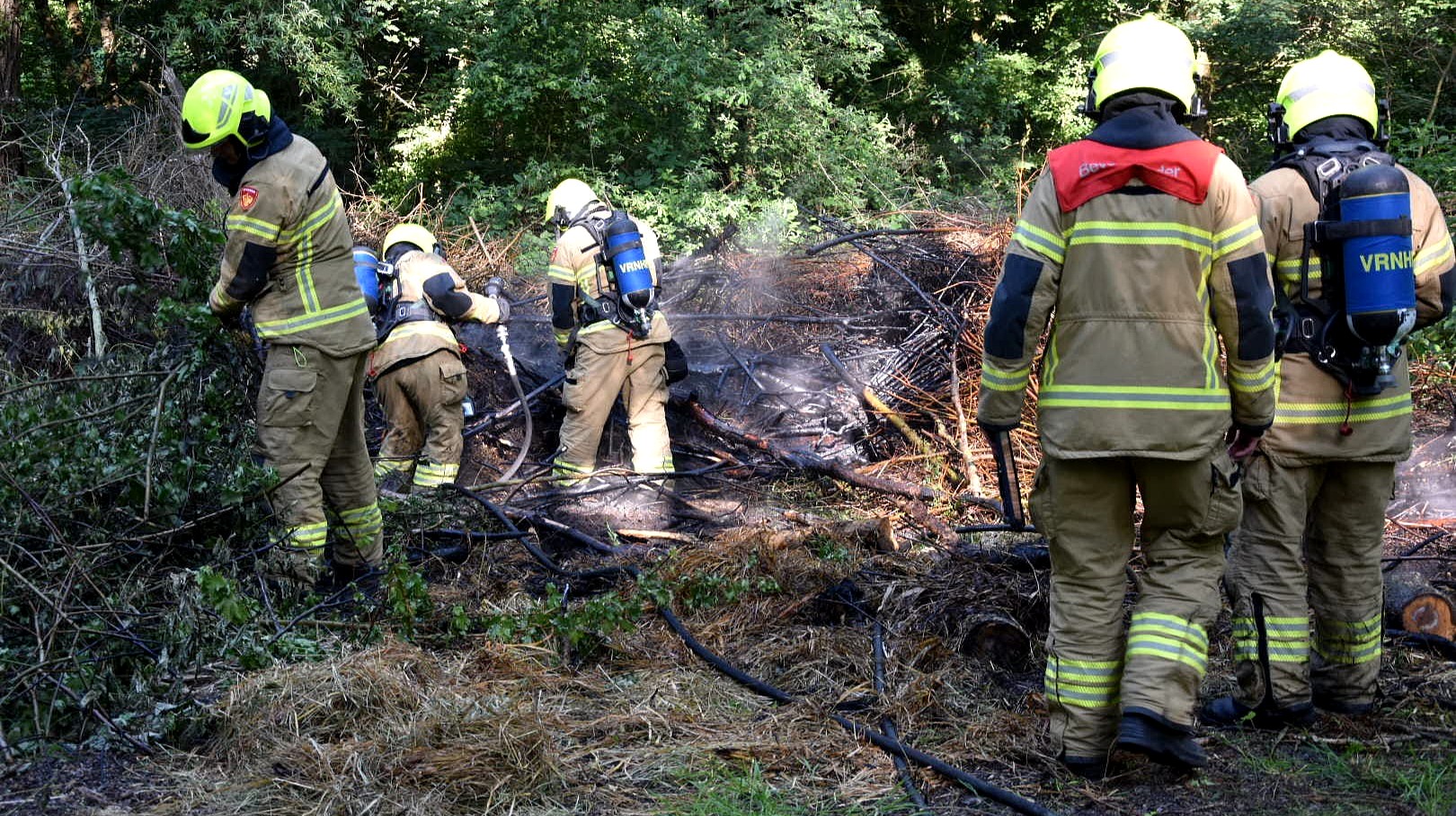 Brandweerlieden blussen een smeulende bosbrand, gekleed in volledige beschermende uitrusting, in een bosrijke omgeving.