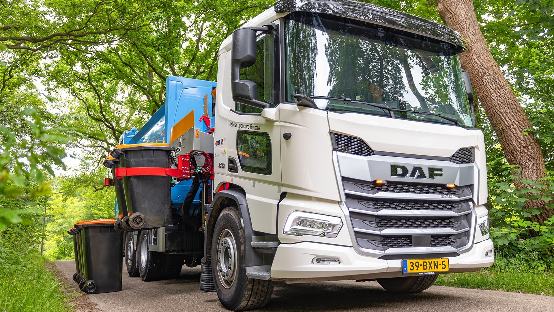 Vuilniswagen van DAF met grijparm die vuilnisbakken optilt, geparkeerd op een smalle weg met bomen aan beide zijden.