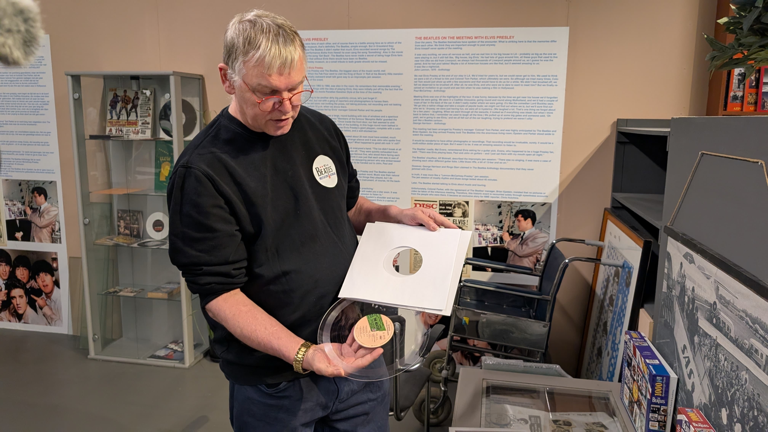 Een man houdt een vinylplaat in een kartonnen hoes vast in een ruimte met tentoonstellingspanelen en Beatles-gerelateerde memorabilia.
