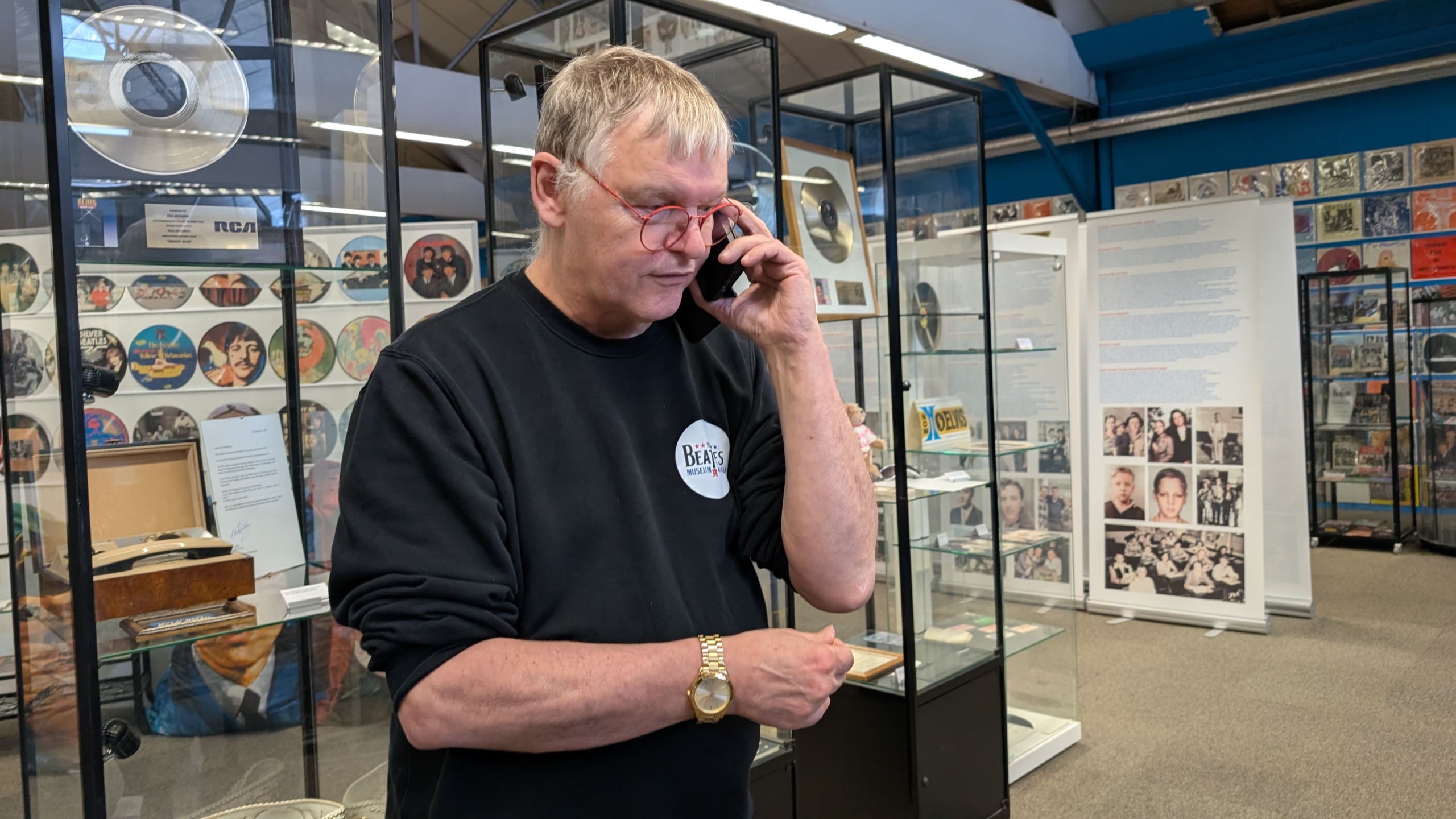Man met een zwart T-shirt en een bril met rode montuur praat aan de telefoon in een museum vol met Beatles-memorabilia, waaronder een glazen vitrinekast en foto's.