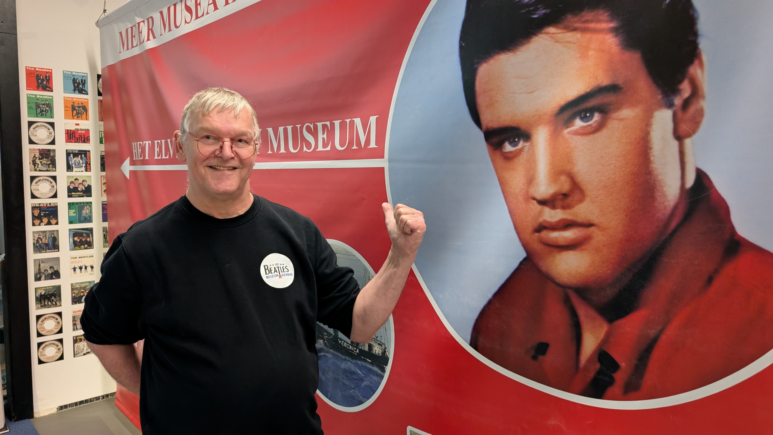 Een man met een zwarte trui en rode bril wijst naar een grote foto van een man in een rode jas op een museumdisplay met de tekst "Het Elvis Museum". Op de achtergrond zijn afbeeldingen van Beatles-albums te zien.