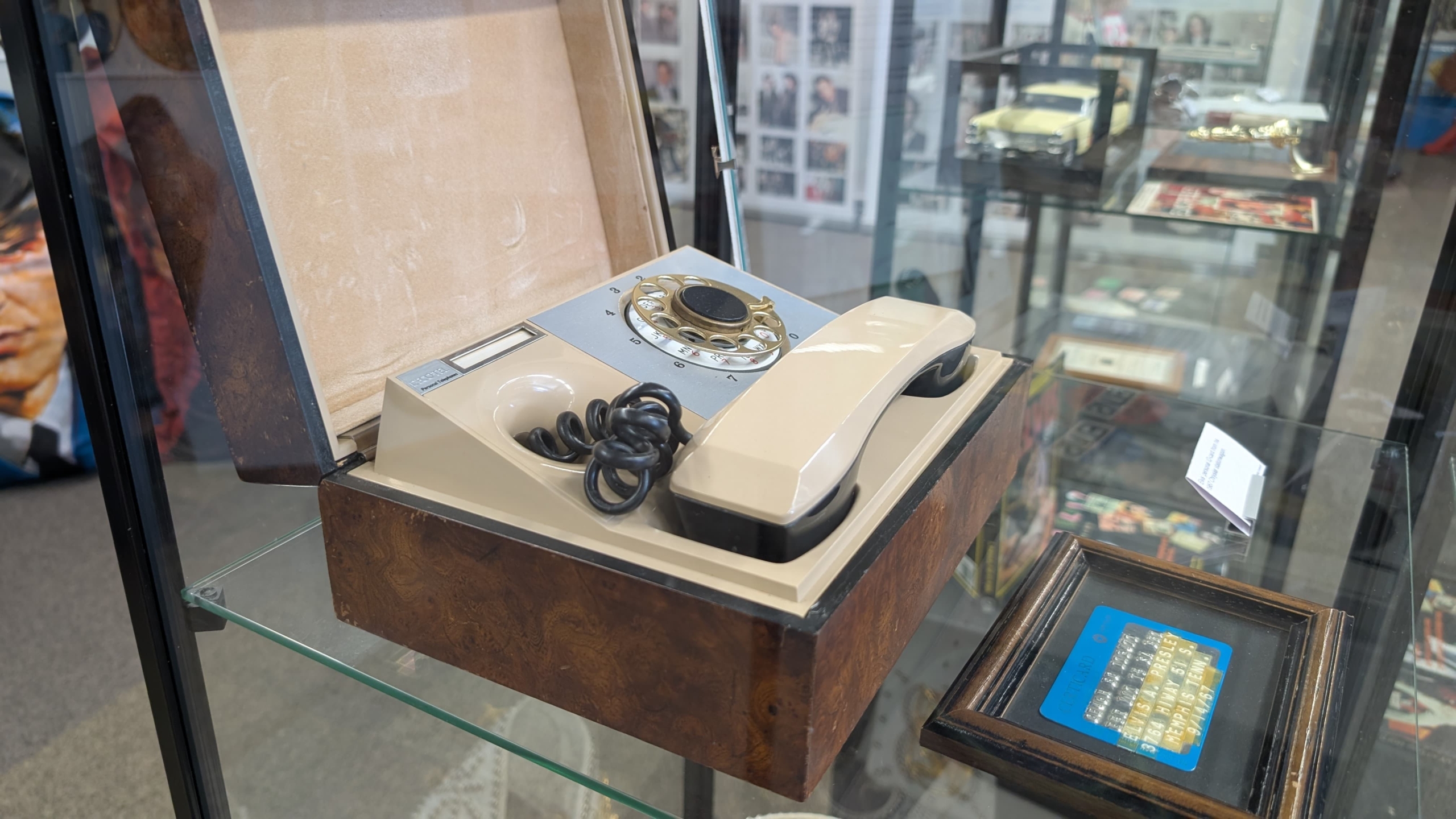 Een antieke beige draaischijftelefoon in een glazen vitrine, omgeven door andere memorabilia in een museumachtige setting.