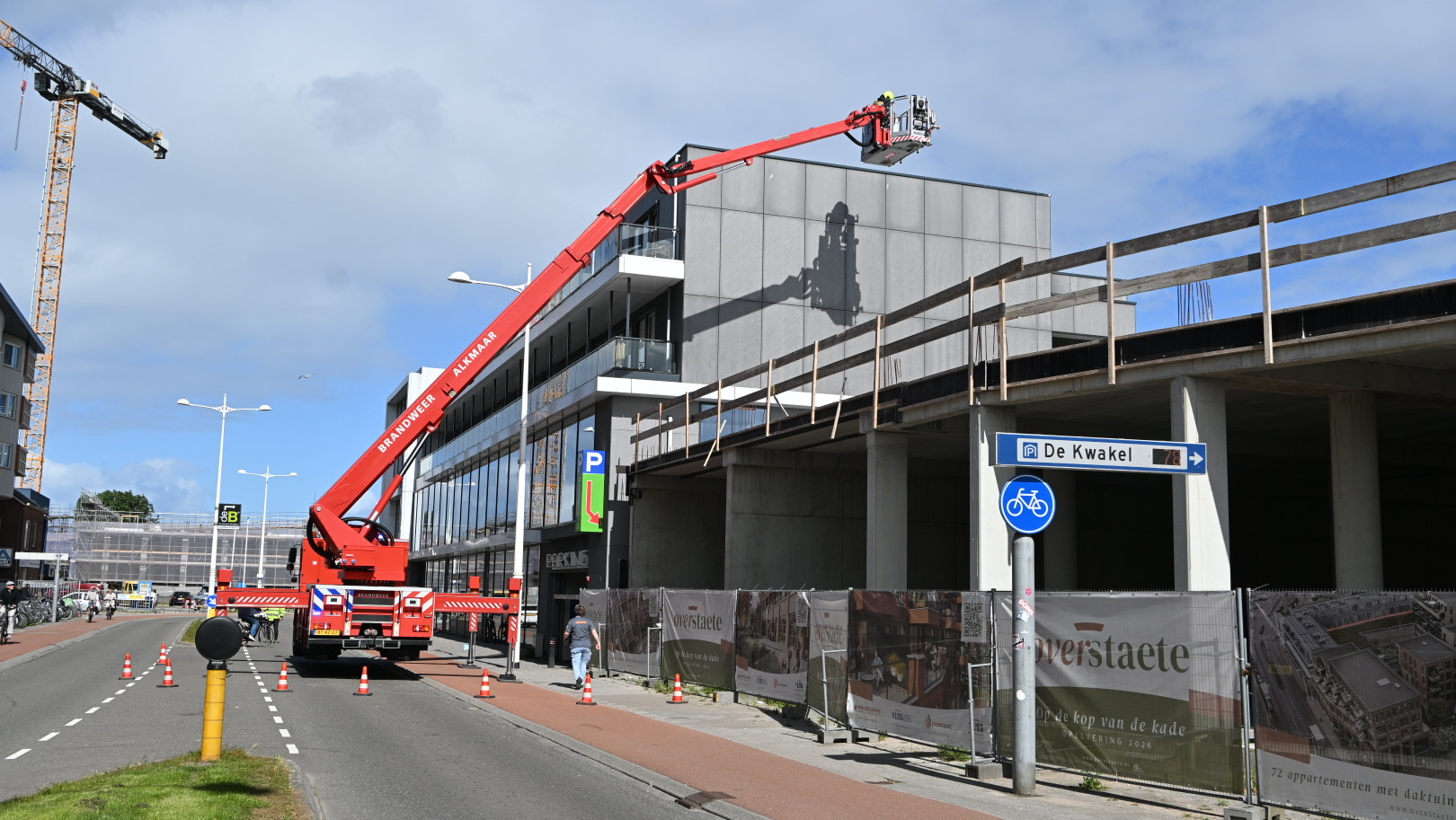 Een rode brandweerwagen met een uitschuifbare ladder voert werkzaamheden uit naast een gebouw in de stad Alkmaar, met een bouwkraan op de achtergrond en een dekkleed met informatie over nieuwe appartementen aan de zijkant van het gebouw.