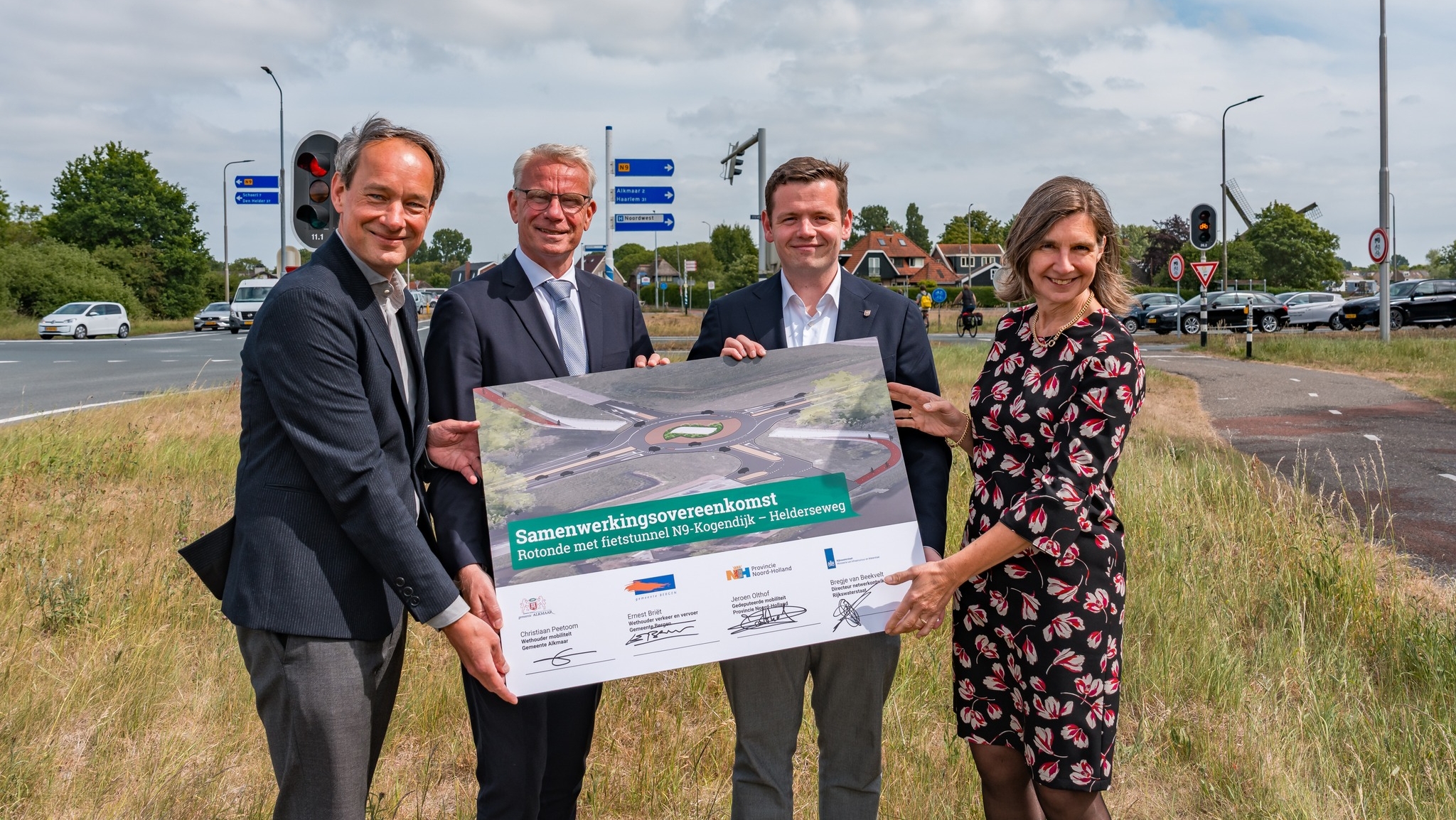 Vier mensen staan buiten langs een weg en houden gezamenlijk een groot bord vast waarop de tekst 'Samenwerkingsovereenkomst Rotonde met fietstunnel N9-Kogendijk - Heldersweg' staat, met een afbeelding van de geplande rotonde.