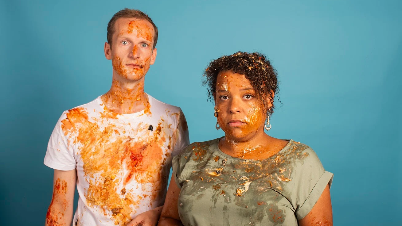 Twee mensen bedekt met voedselresten staan tegen een blauwe achtergrond, de ene persoon draagt een witte T-shirt en de andere een groene top.