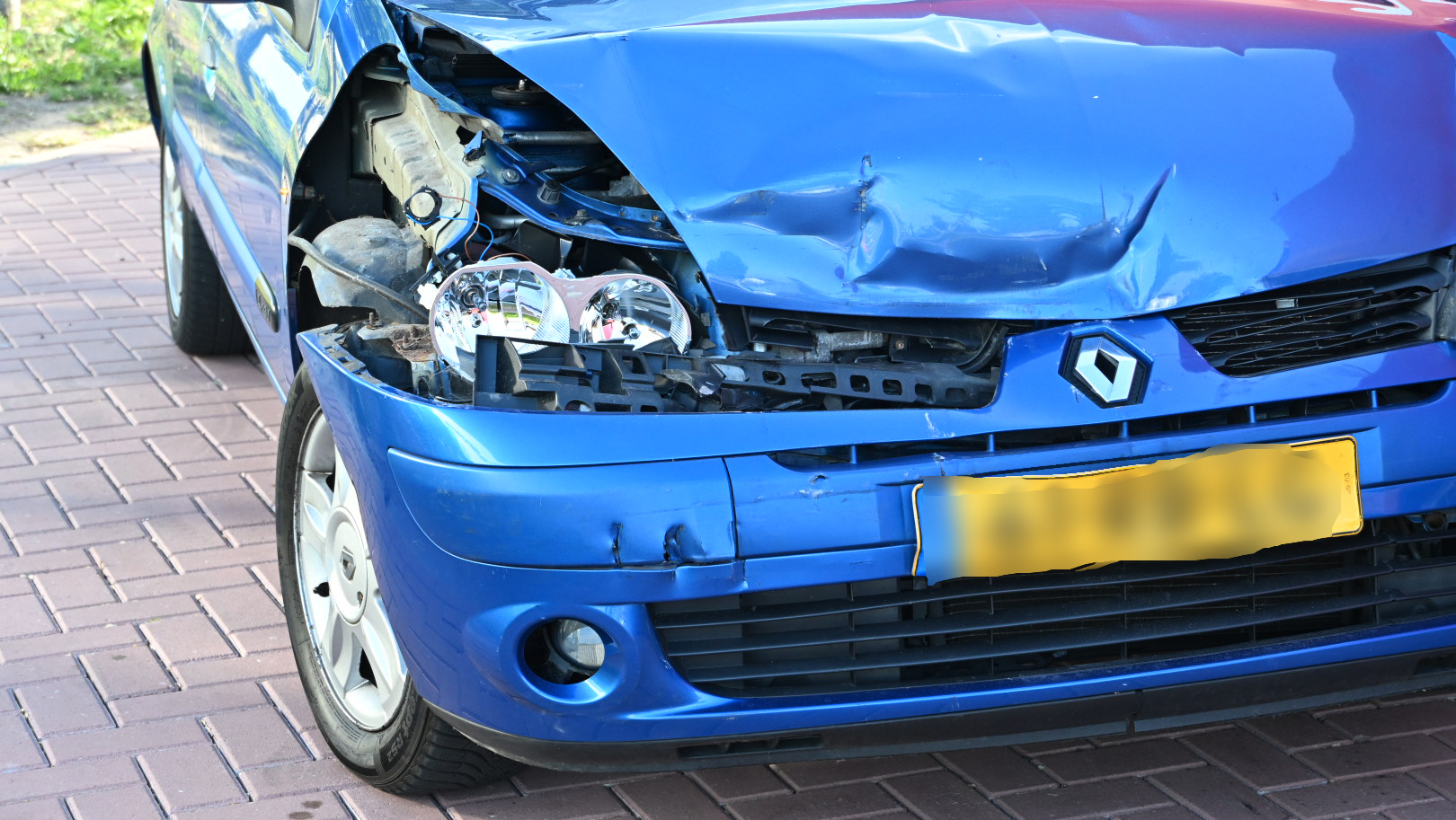 Blauwe auto met aanzienlijke schade aan de voorkant, met een gedeukt en gebroken motorkap en koplampen, geparkeerd op een bestrate oprit.