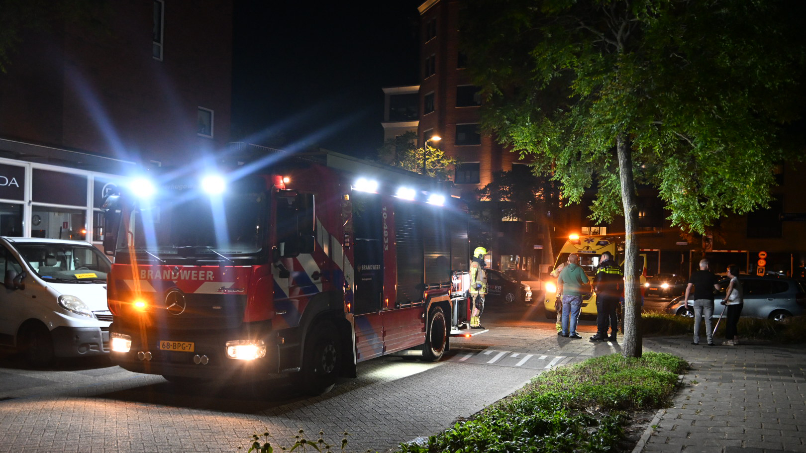 Een brandweerwagen met felle lichten staat 's nachts geparkeerd op een straat naast een gebouw, omgeven door enkele mensen, waaronder een brandweerman en een politieagent.