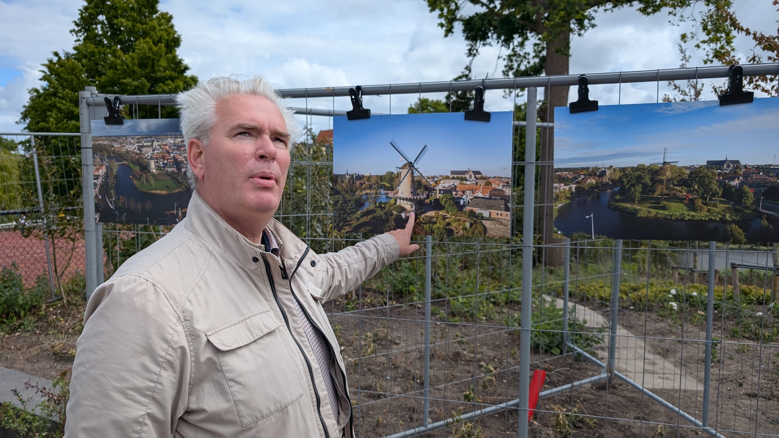 Een man wijst naar een foto van een windmolen, tentoongesteld op een hek. Er zijn meerdere foto's te zien tegen een achtergrond van bomen en een heldere lucht.