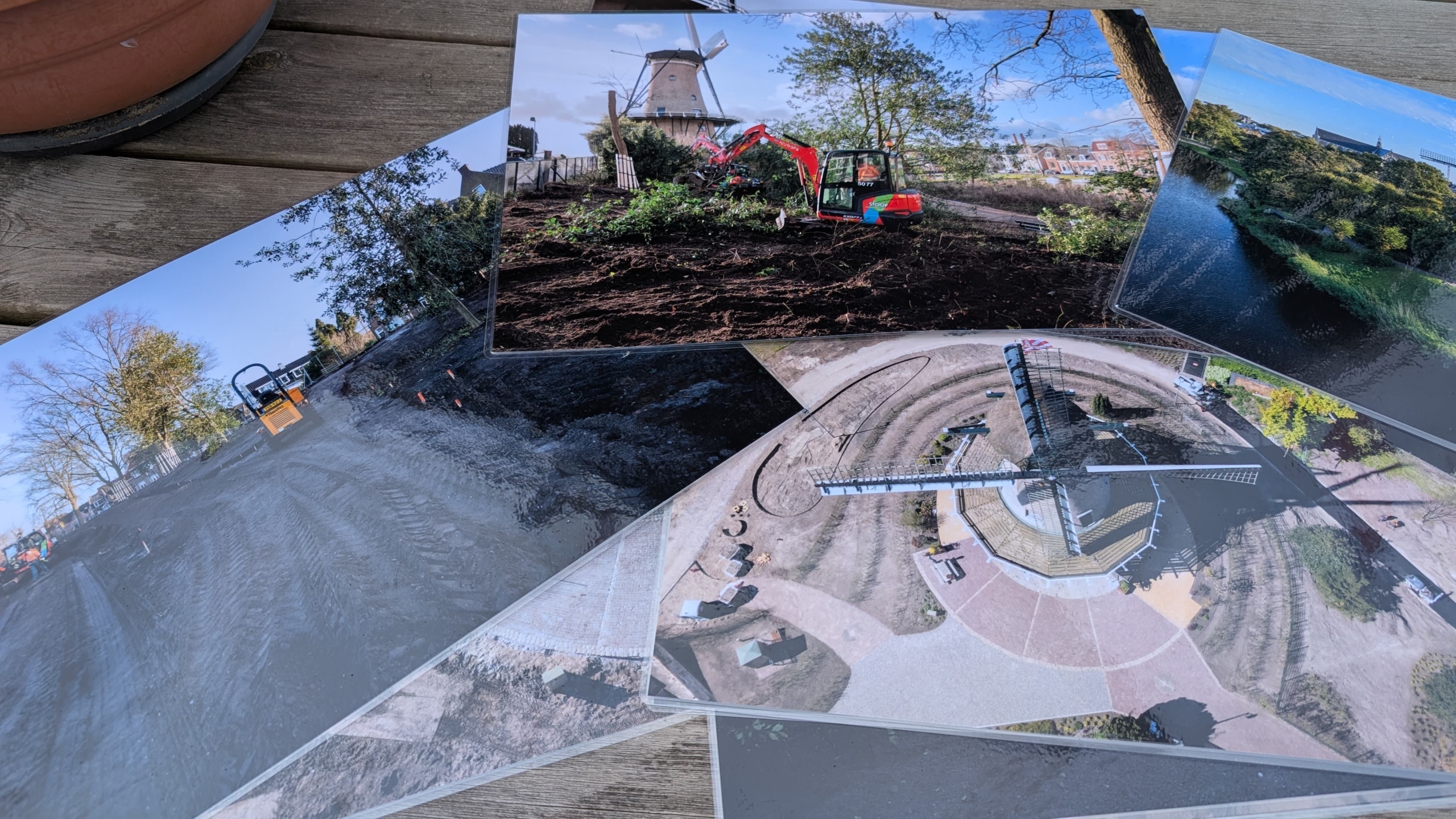 Een verzameling geprinte foto's van een bouw- of landschapsproject op een houten tafel. De bovenste foto toont een rode graafmachine bij een oude windmolen, terwijl andere foto's graafwerkzaamheden en een hoog uitzicht op de omgeving tonen.