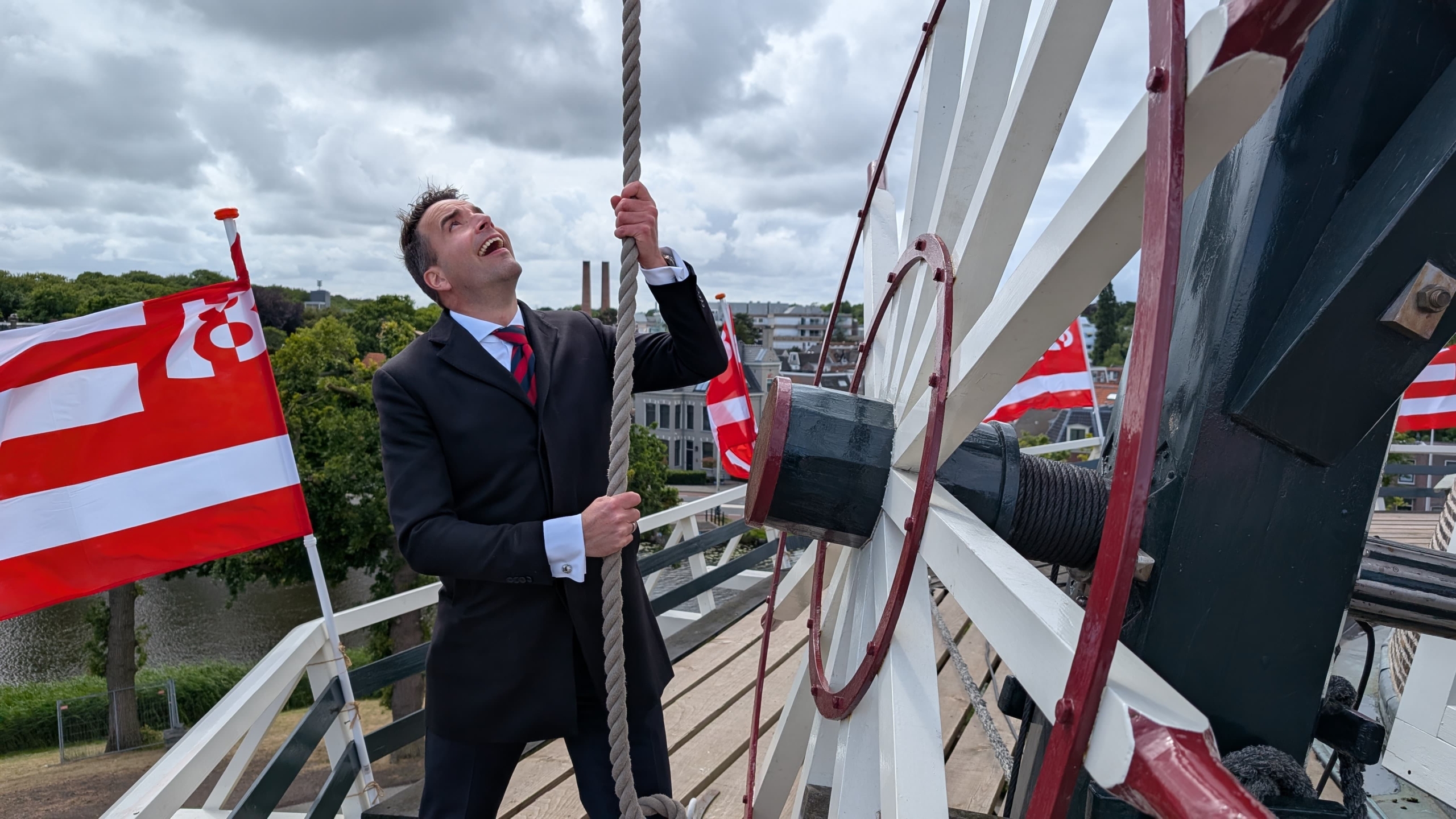 Man in a suit pulling a rope on a traditional Dutch windmill with red and white flags in the background.
