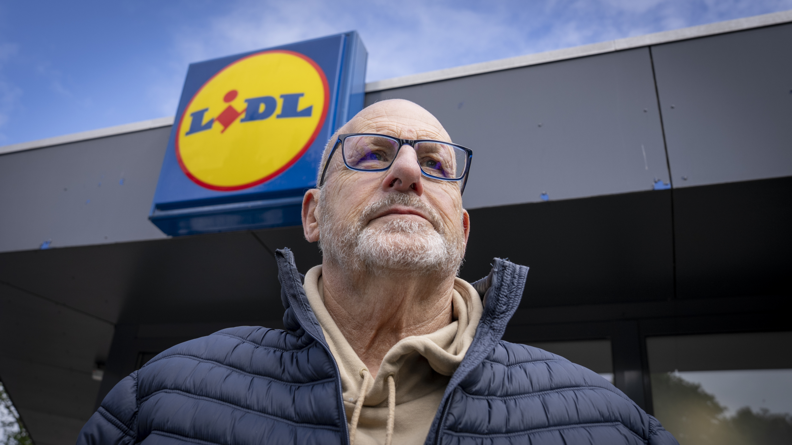 Een oudere man met een bril kijkt omhoog terwijl hij voor een Lidl-winkel staat, met het logo van de supermarkt op de achtergrond.