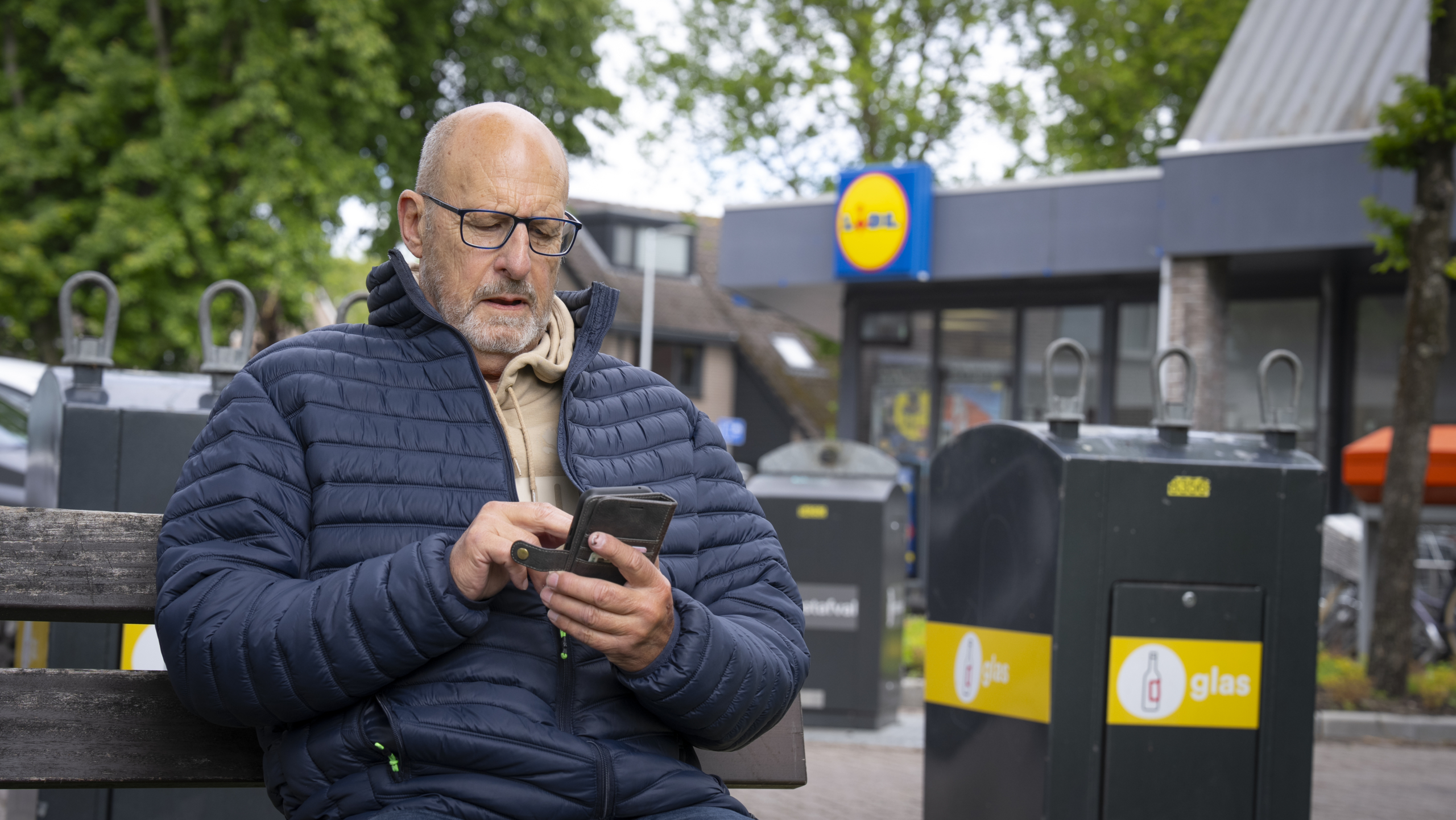 Een man met een bril en een winterjas zit op een bank en kijkt op zijn smartphone, met op de achtergrond een Lidl supermarkt en glascontainers.