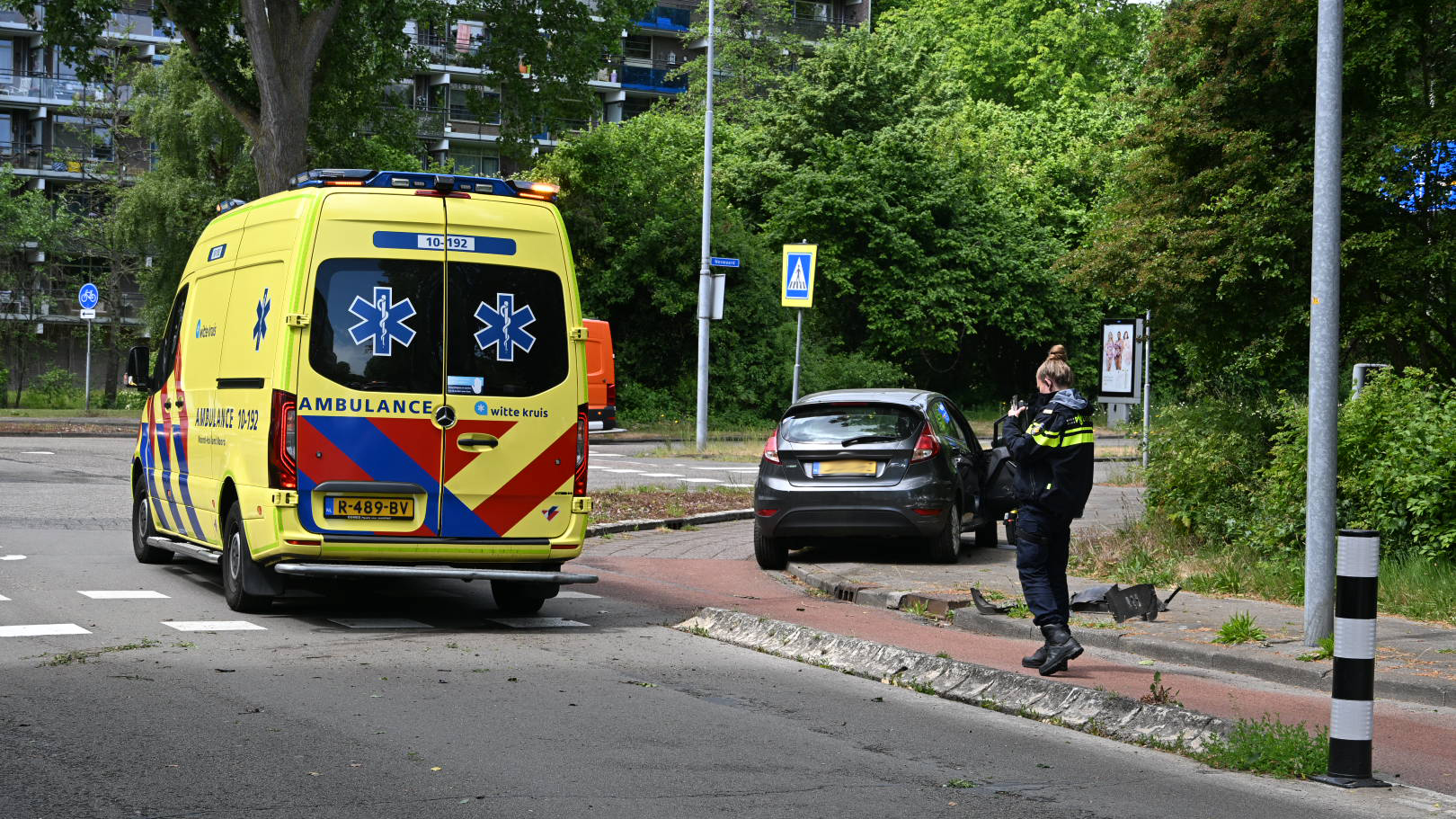 Een gele ambulance met blauwe en rode strepen staat geparkeerd op een straat in een groene omgeving, naast een beschadigde zwarte auto en een politiemedewerker die foto's maakt van de situatie.