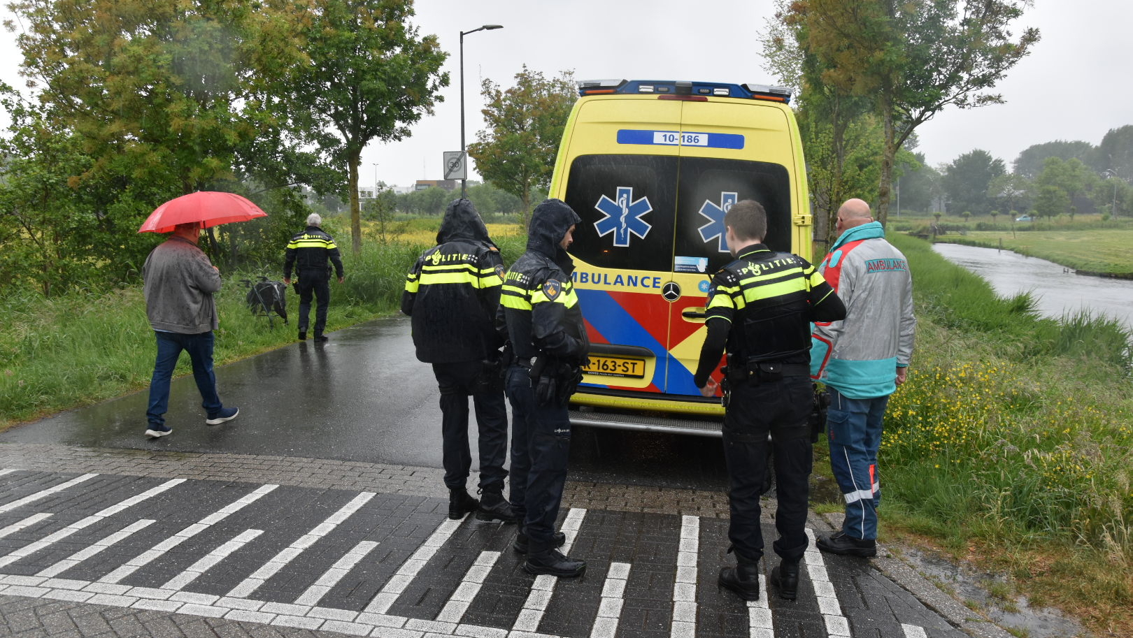 Politieagenten en een ambulance-medewerker staan in de regen naast een ambulance bij een landelijk weggetje; een omstander met rode paraplu loopt weg.