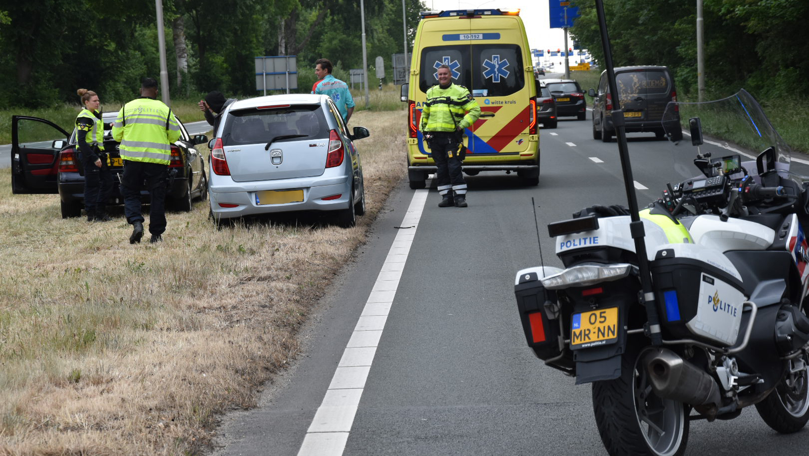 Politie en een ambulance bij een auto aan de kant van de weg na een incident, met agenten en betrokkenen aanwezig.