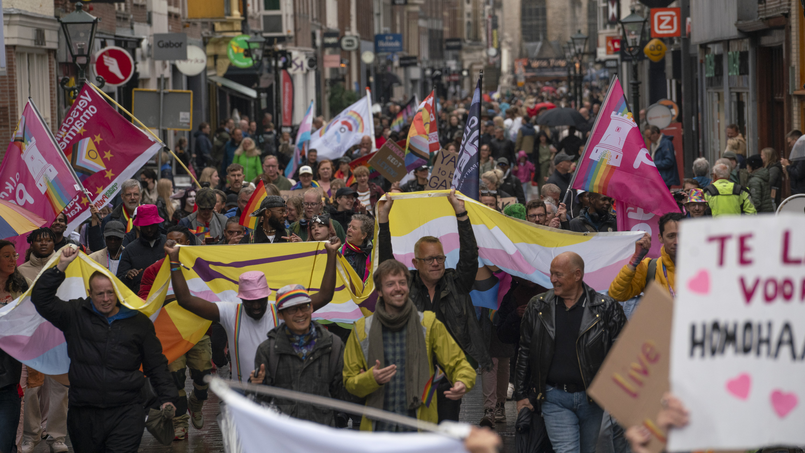 Een groep mensen loopt mee in een kleurrijke parade op straat, met regenboogvlaggen en borden met ondersteunende boodschappen voor de LGBTQ+ gemeenschap.