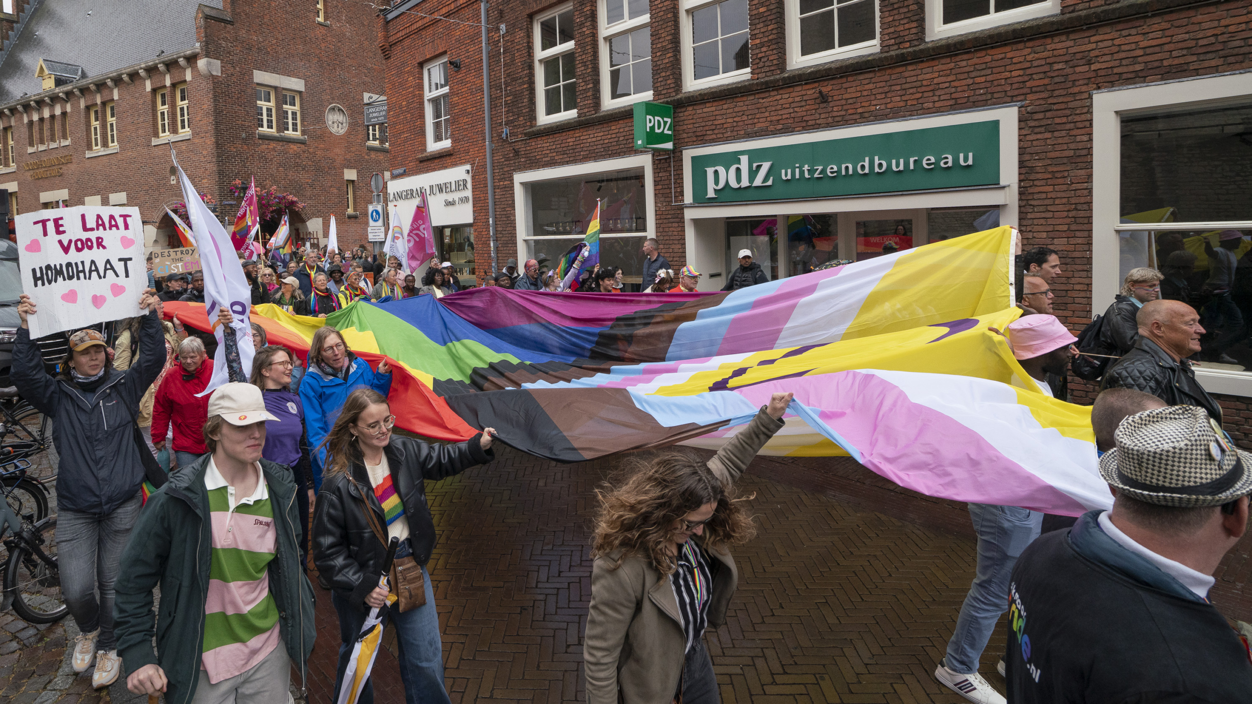 Een groep mensen loopt mee in een pride parade, waarbij ze een grote regenboogvlag vasthouden. Een van de deelnemers houdt een bord vast met de tekst 'Te laat voor homdhaat'. Op de achtergrond zijn historische gebouwen en een uitzendbureau zichtbaar.