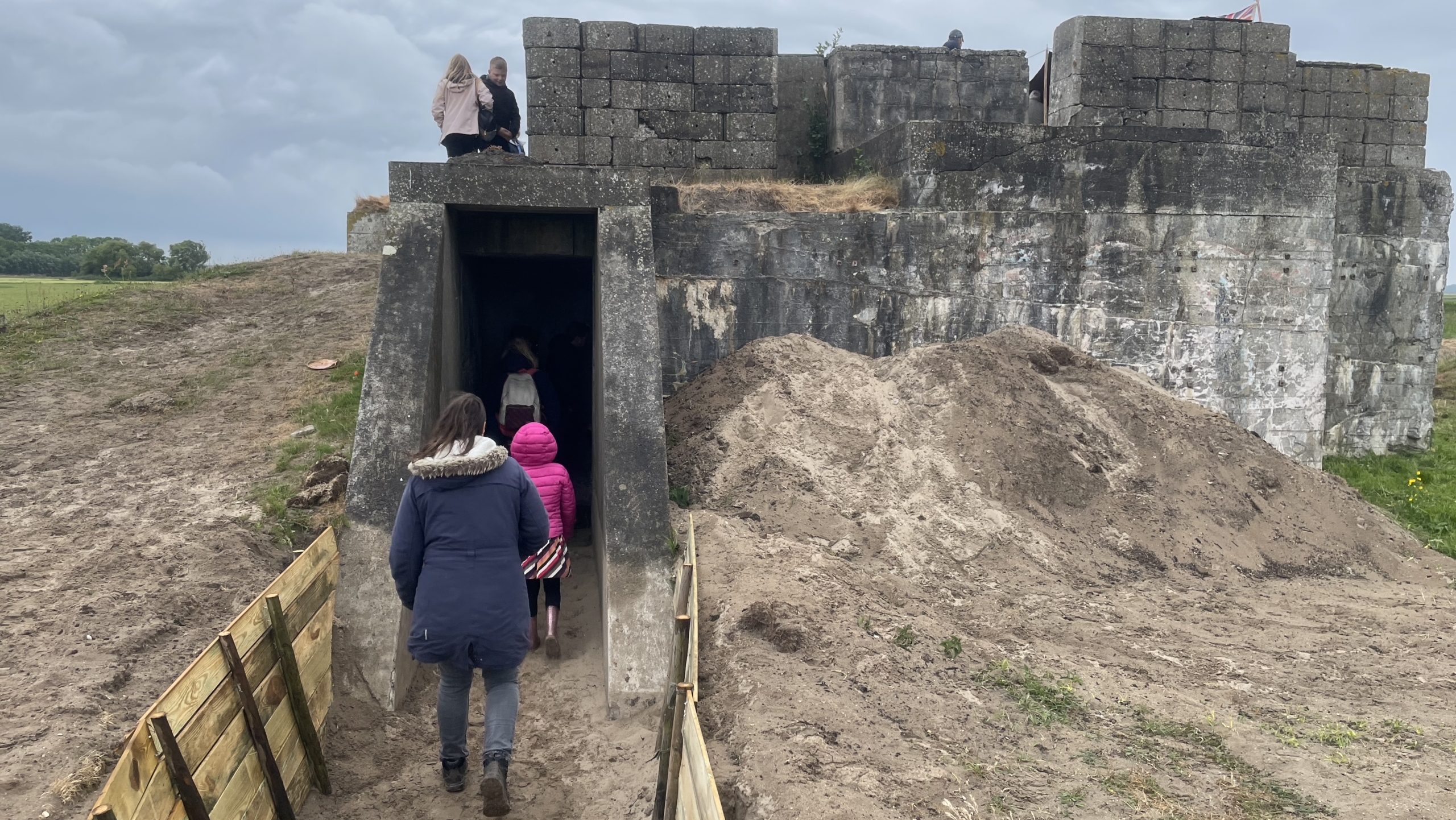 Mensen lopen een betonnen bunker binnen, terwijl anderen bovenop zitten; de bunker ligt in een grasrijke omgeving met zandige aarde en gedeeltlijke houten bekisting langs het pad.