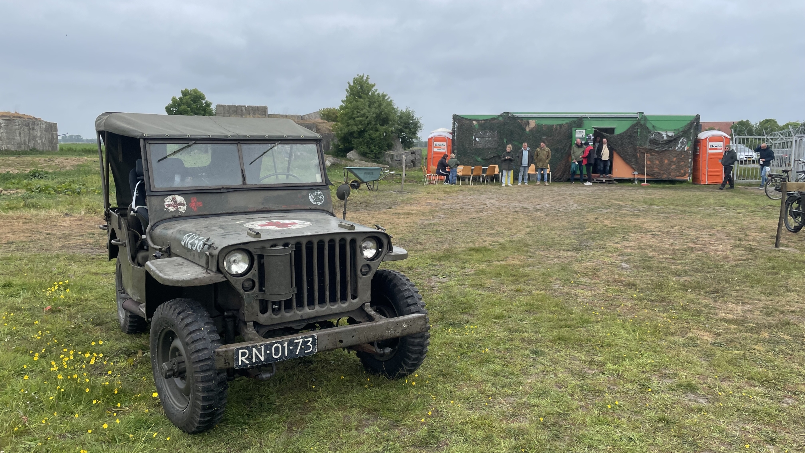 Een oude legerjeep staat geparkeerd op een grasveld, met een klein gebouw en enkele mensen op de achtergrond. De lucht is bewolkt en er zijn veldbloemen op de voorgrond.
