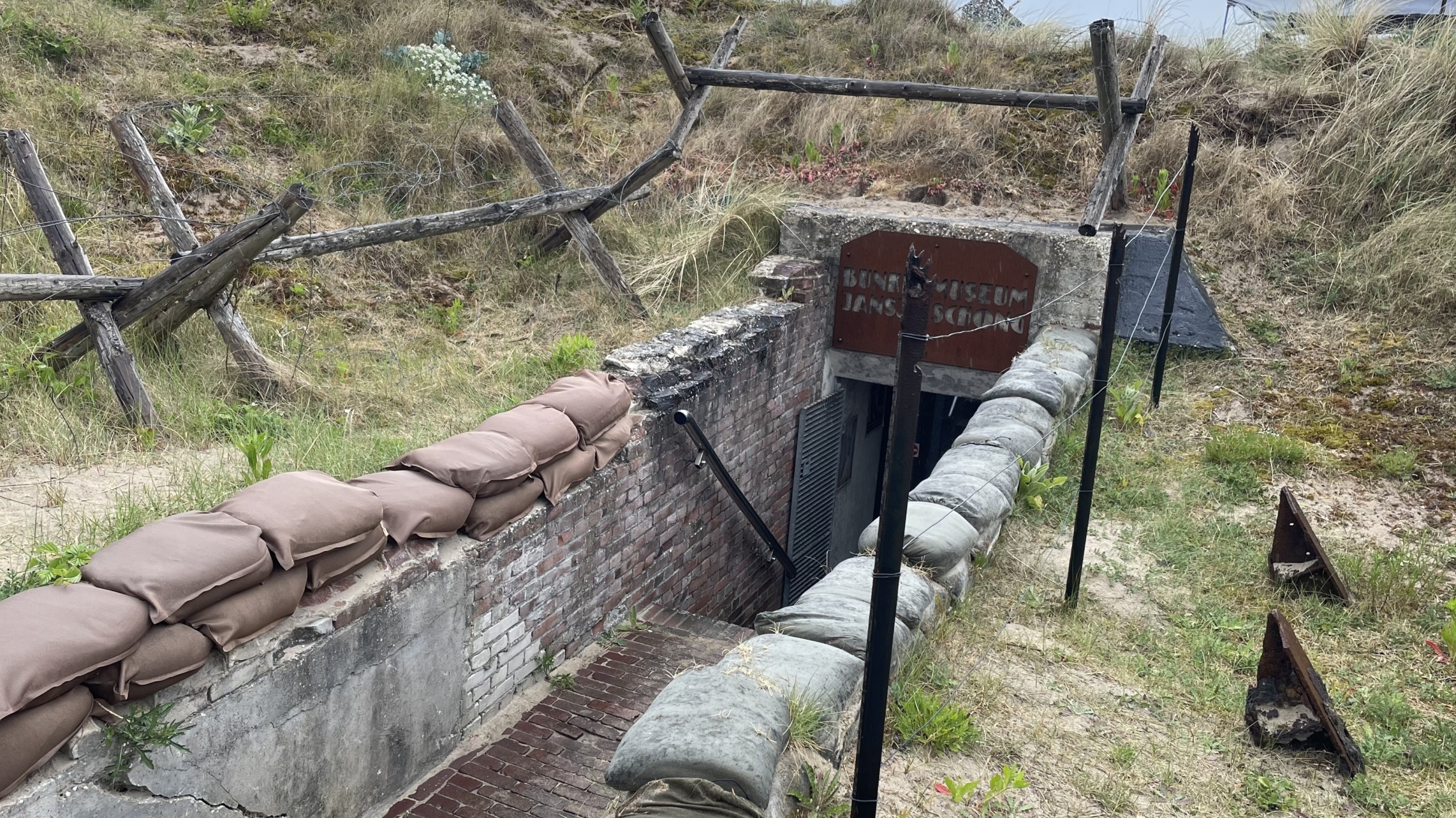 Ingang van een bunker met zandzakken rondom, omgeven door gras en houten obstakels, met een bord dat "Bunkermuseum Jans Schouten" aangeeft.