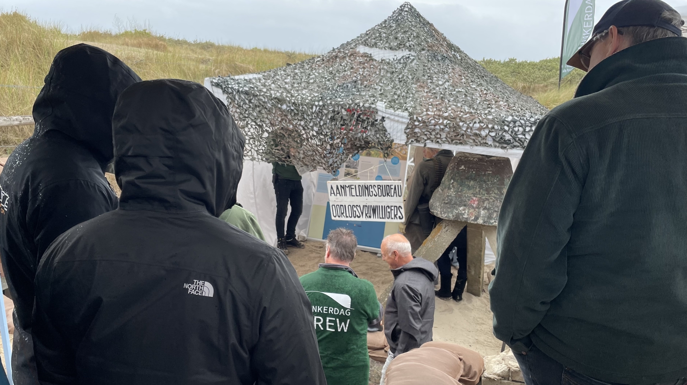 Groep mensen in regenjacks verzameld bij een tent met een camouflage net, met een bord voor 'Aanmeldingsbureau Oorlogs Vrijwilligers' in een duinachtige omgeving.