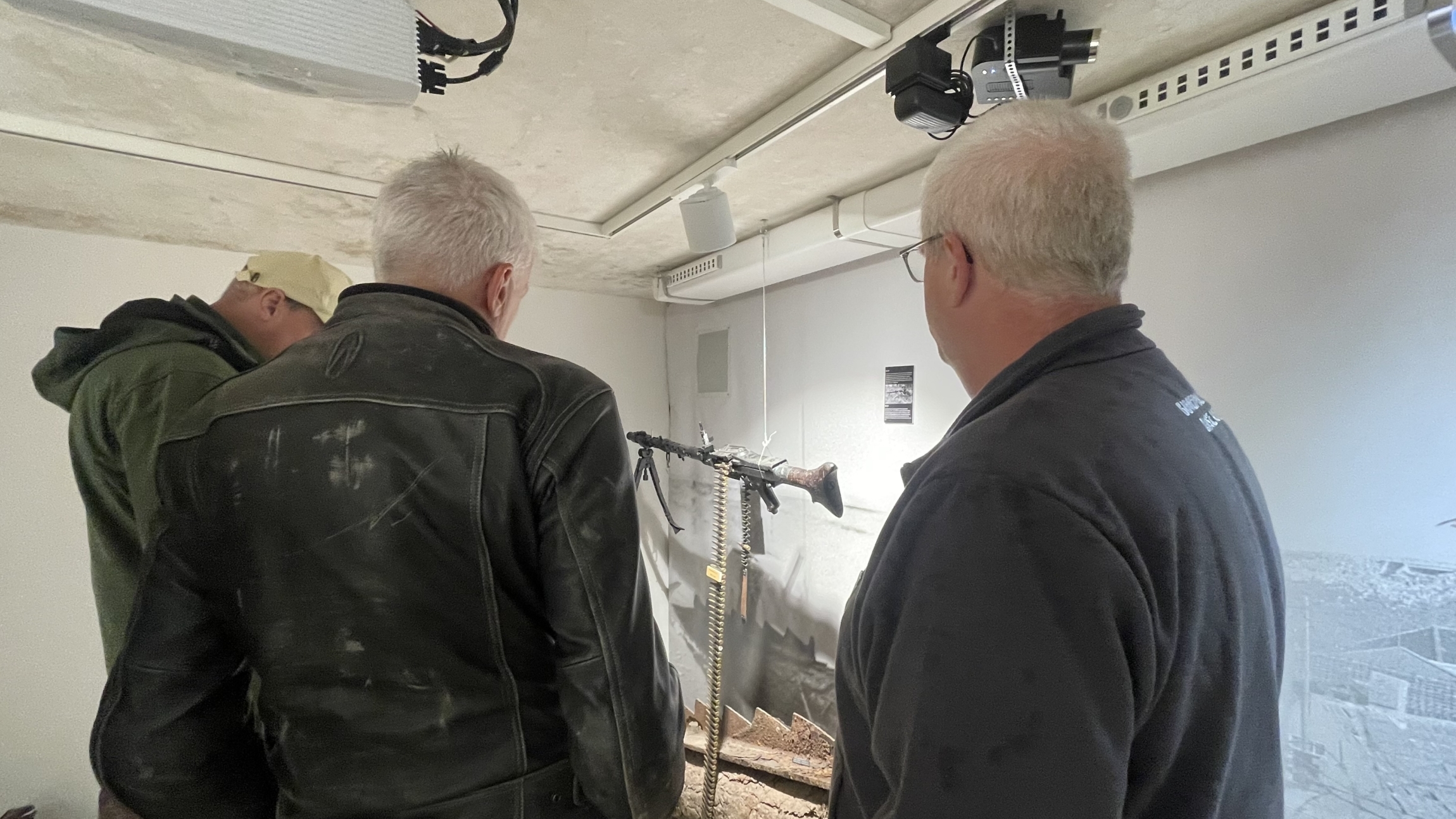 Drie mannen bekijken een opstelling van een machinegeweer in een museumachtig interieur, met een plafondprojector boven hen.