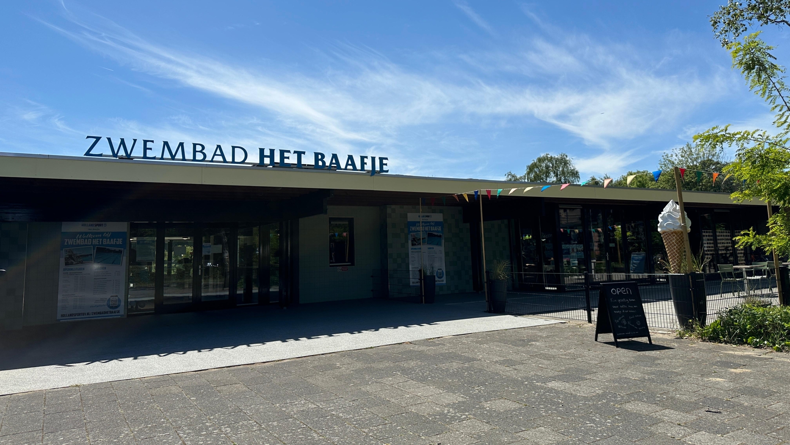 Ingang van Zwembad Het Baafje, met boven de ingang een groot bord met de naam van het zwembad en links een groot ijsje als decoratie op een zonnige dag.