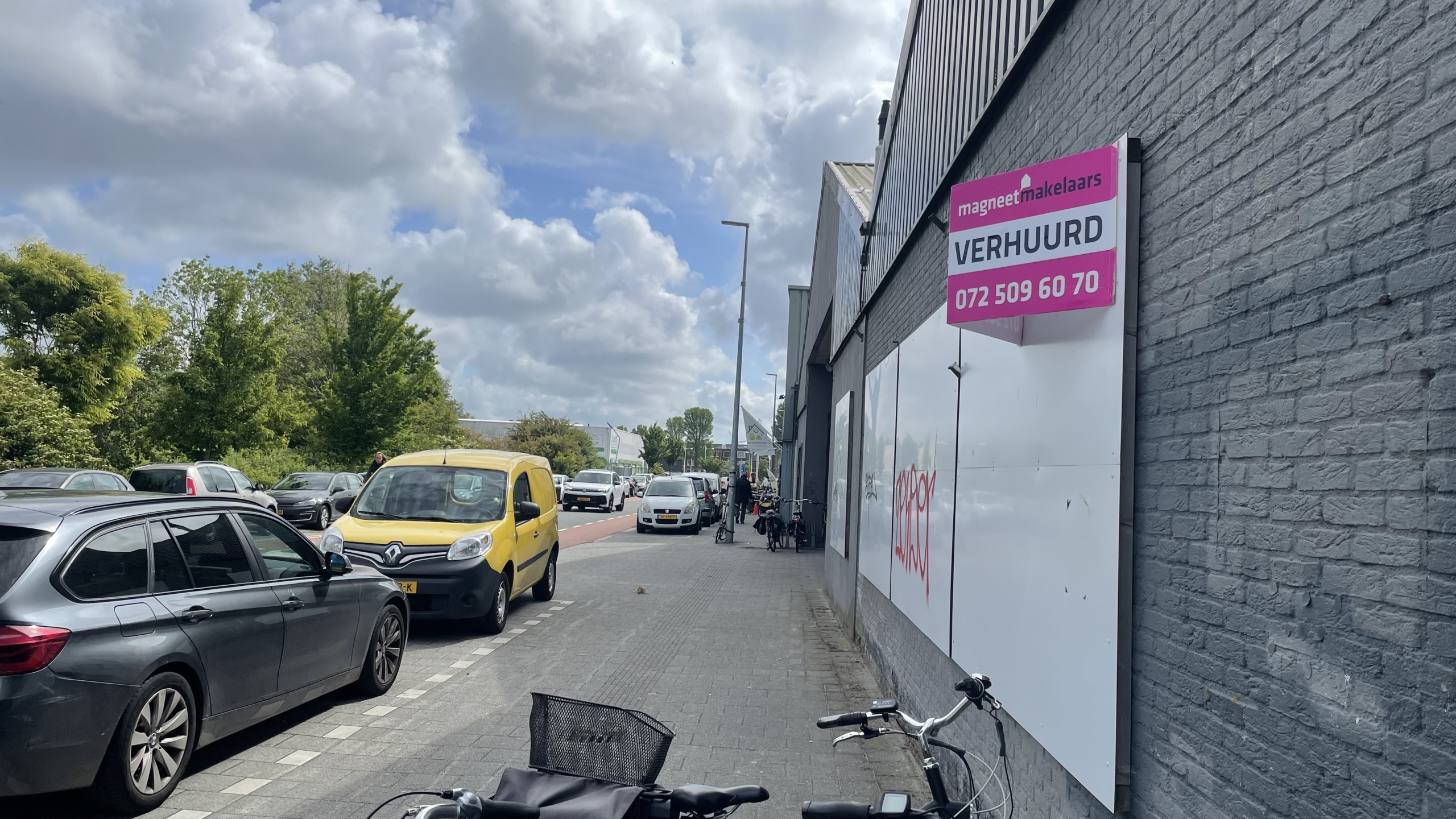Straatbeeld met verschillende auto's geparkeerd langs de weg naast een grijs gebouw. Aan het gebouw hangt een bord van "magneet makelaars" met de tekst "VERHUURD" en een telefoonnummer. Er zijn fietsen geparkeerd langs de muur en de lucht is deels bewolkt.