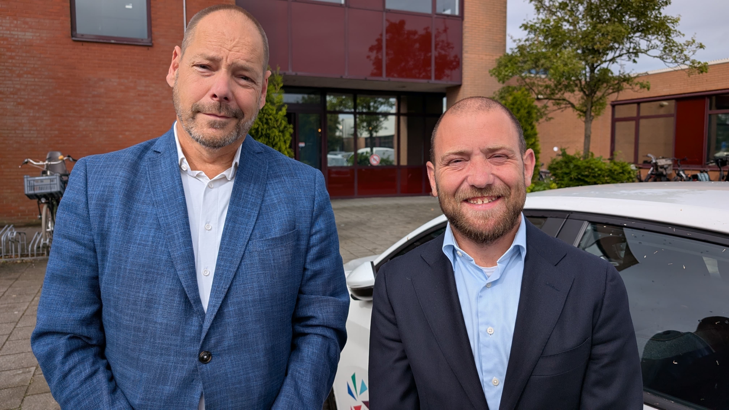 Twee mannen in pakken poseren voor een gebouw met rode bakstenen en grote ramen; op de achtergrond staat een fiets naast een auto.