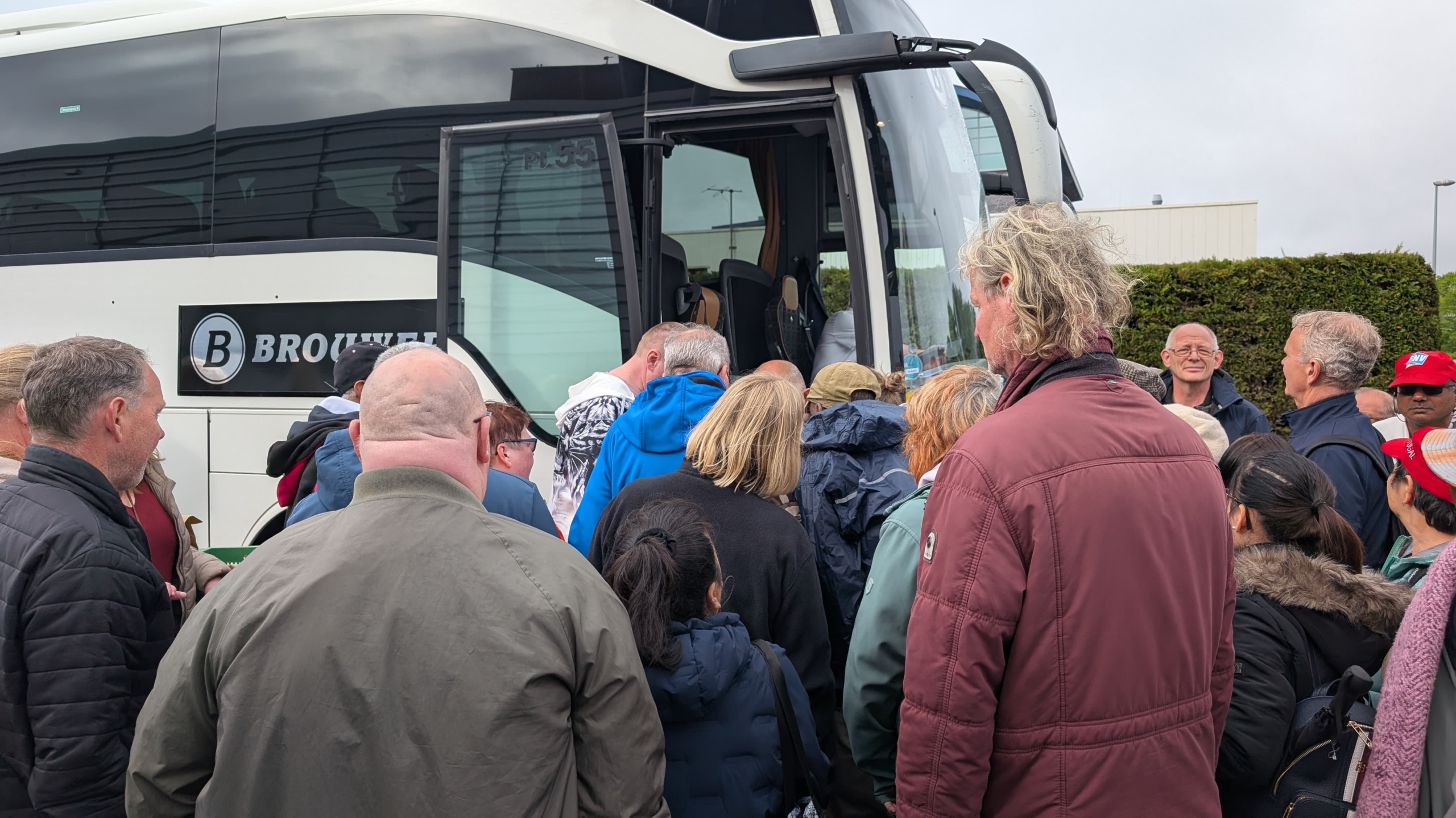 Een groep mensen staat in een rij voor de deur van een touringcar met het logo van "Brouwers" erop.