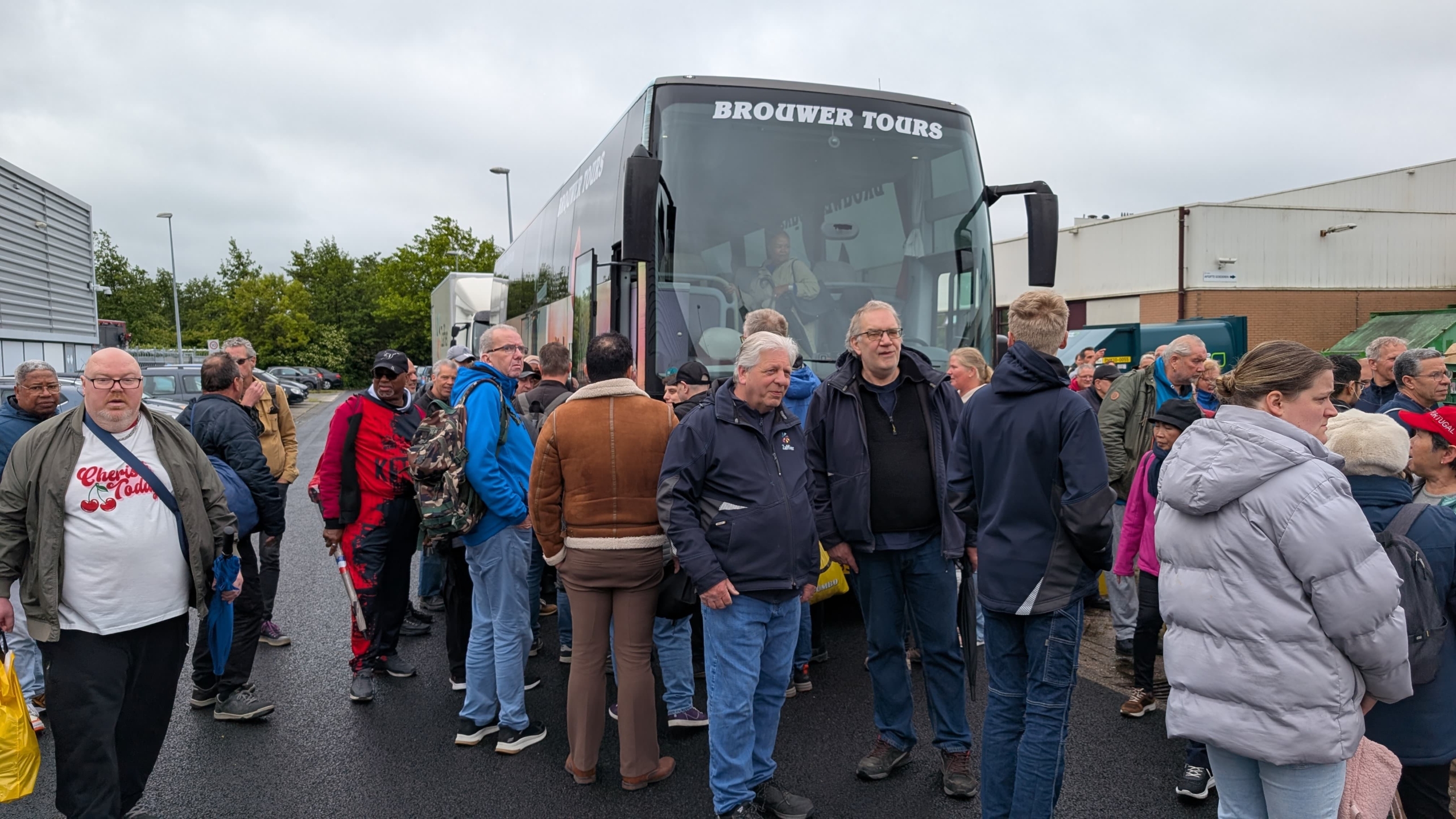 Een groep mensen verzamelt zich bij een bus van Brouwer Tours op een parkeerplaats, met enkele mensen in casual kleding en regenjassen.