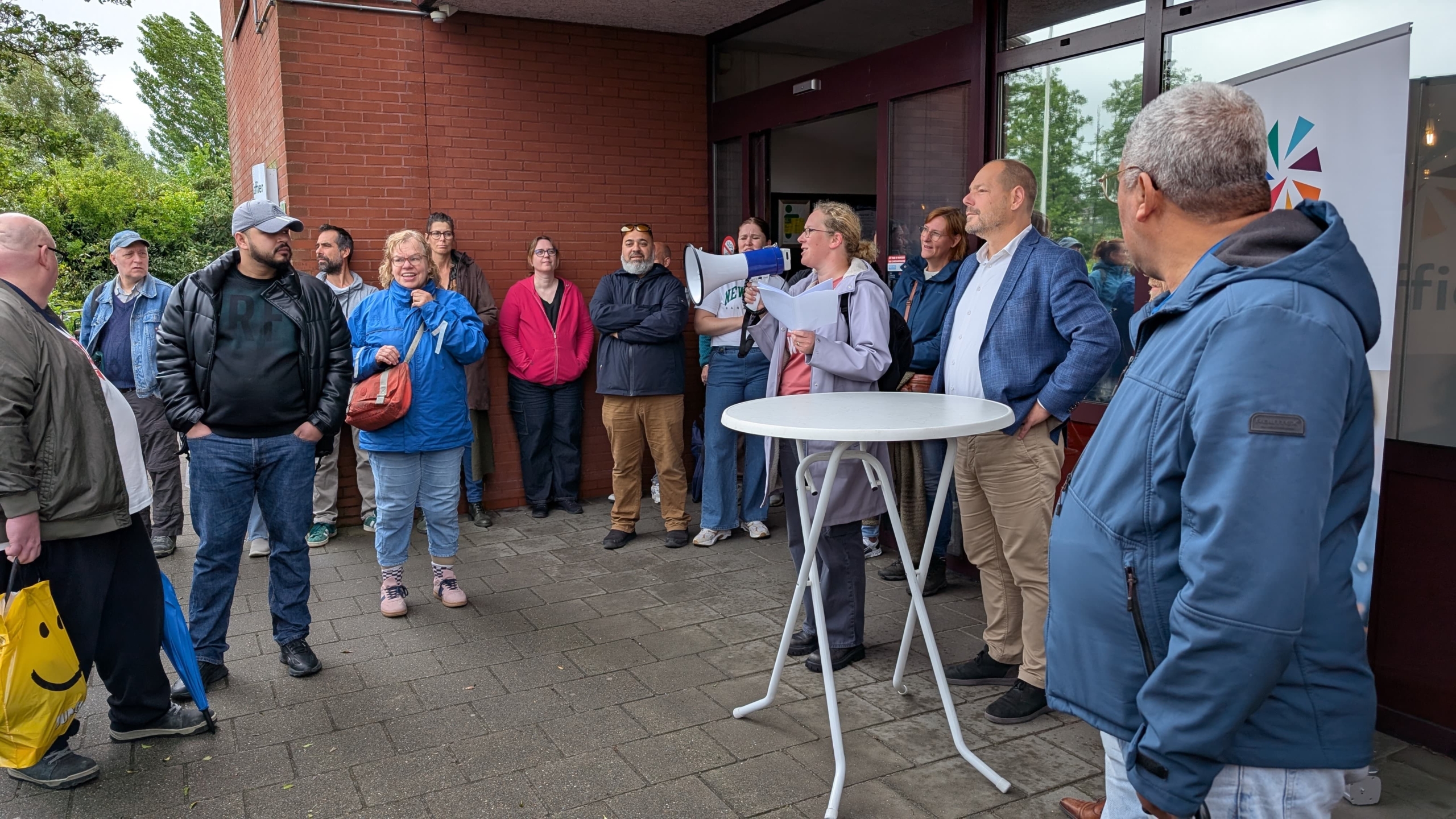 Een groep mensen staat buiten bij een gebouw, terwijl een persoon met een megafoon iets lijkt aan te kondigen. De mensen zijn casual gekleed en luisteren aandachtig. Er staat een ronde tafel op de voorgrond en er is groen gebladerte zichtbaar op de achtergrond.