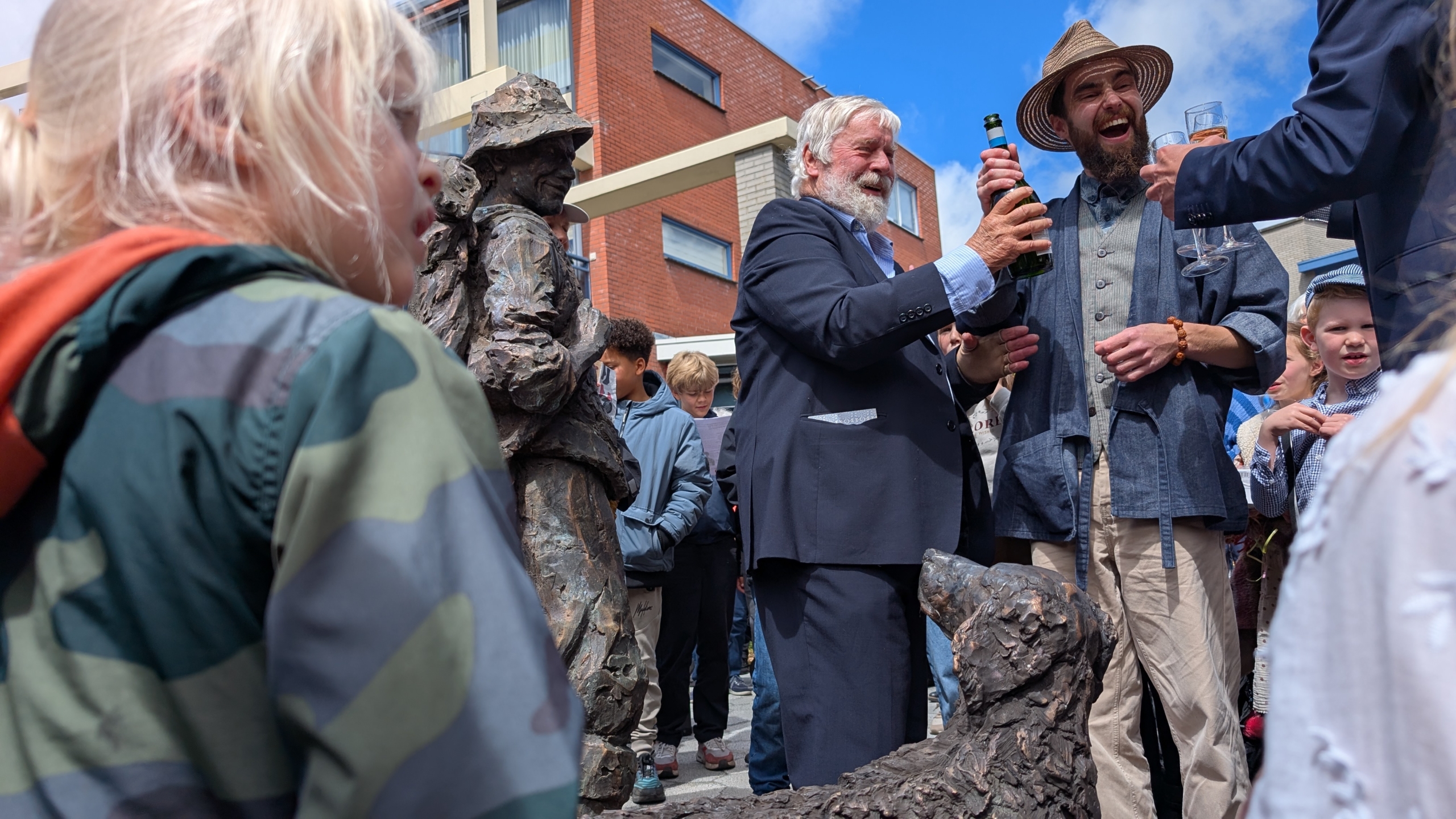 Mensen staan rondom een bronzen standbeeld van een man met een hond, terwijl een oudere man en een andere persoon met hoed een fles openen en toasten. Op de voorgrond een kind in een camouflagejas en meerdere toeschouwers op de achtergrond. Het vindt plaats op een zonnige dag voor een bakstenen gebouw.