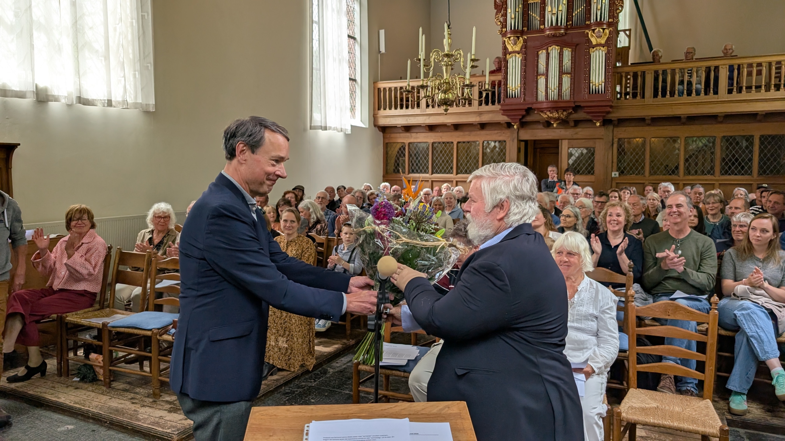 Een man in een donkere jas geeft een boeket bloemen aan een oudere man met grijs haar in een kerk vol toehoorders, terwijl het publiek applaudisseert.
