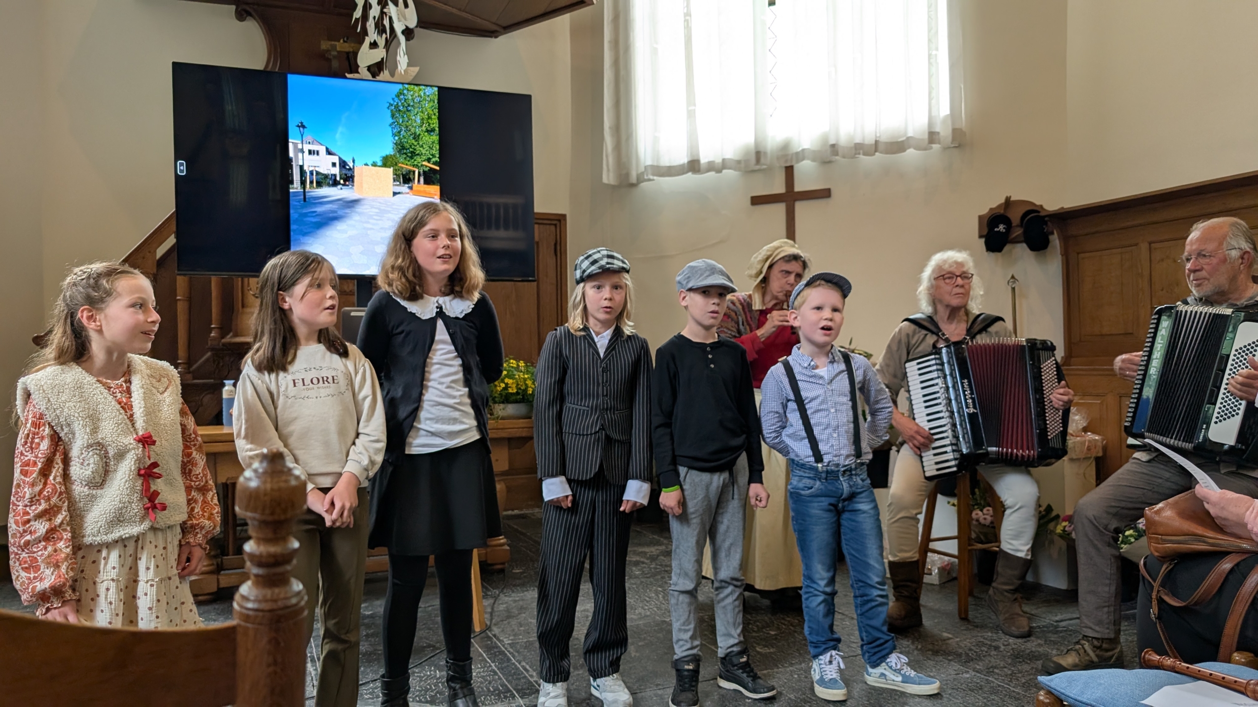Kinderen zingen samen in een kerkzaal terwijl twee volwassenen accordeon spelen; op de achtergrond een scherm met een landschap afbeelding.