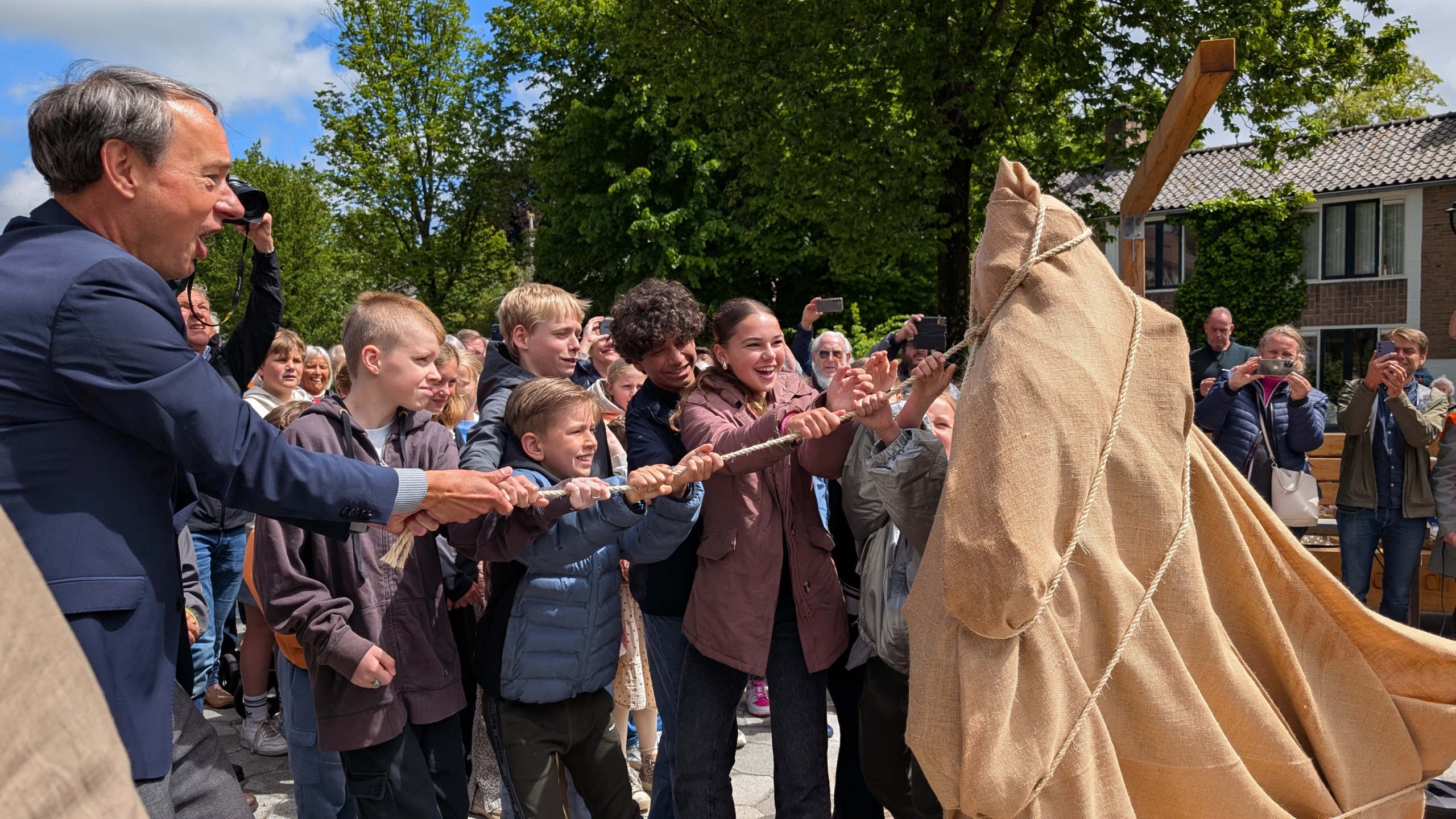 Een groep blije kinderen en een man trekken een touw om een object bedekt met zeildoek te onthullen, omgeven door toeschouwers die foto's maken.