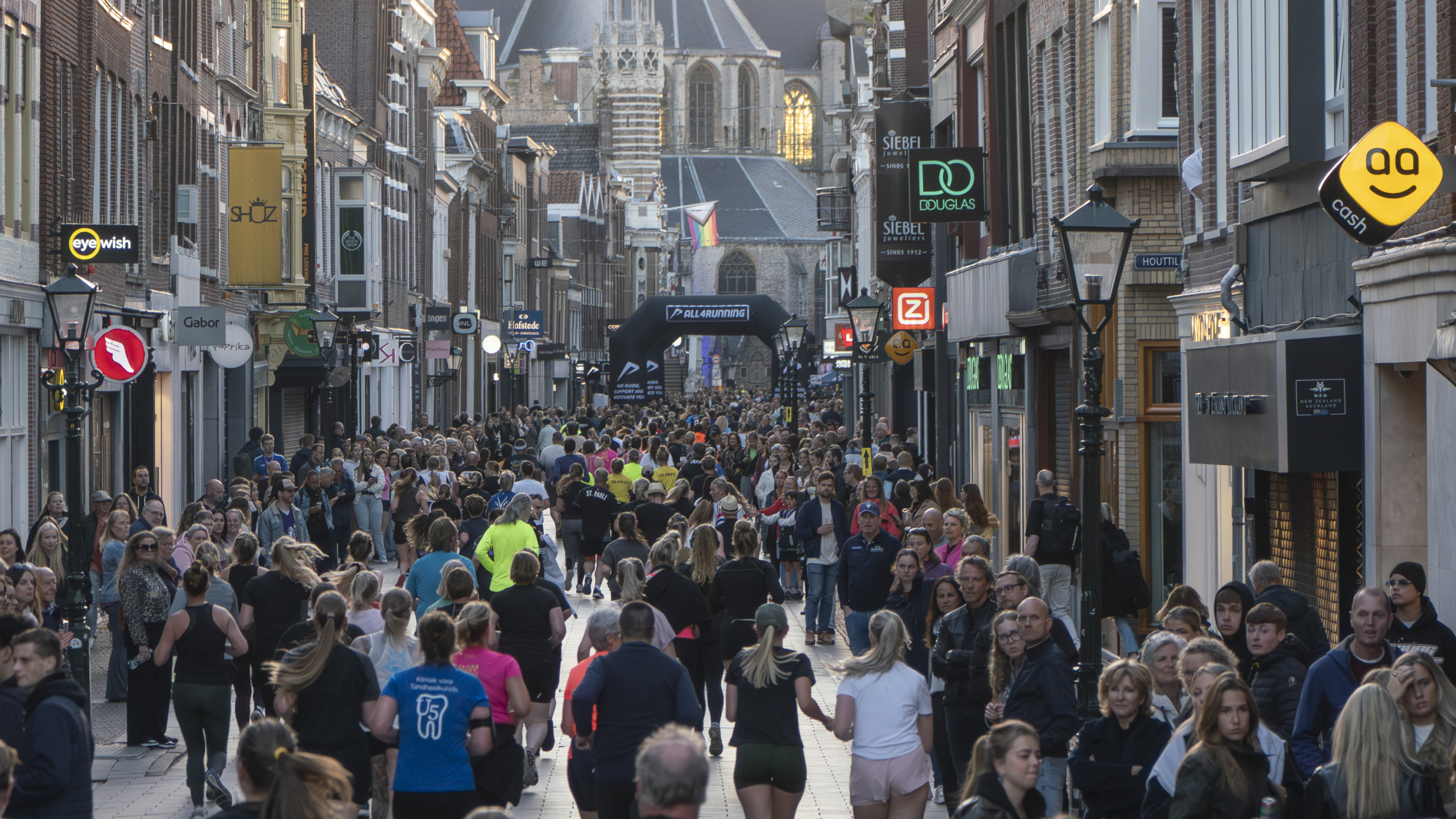 Drukke straat met lopers en toeschouwers tijdens een hardloopevenement in een stadsomgeving met winkels aan beide zijden.
