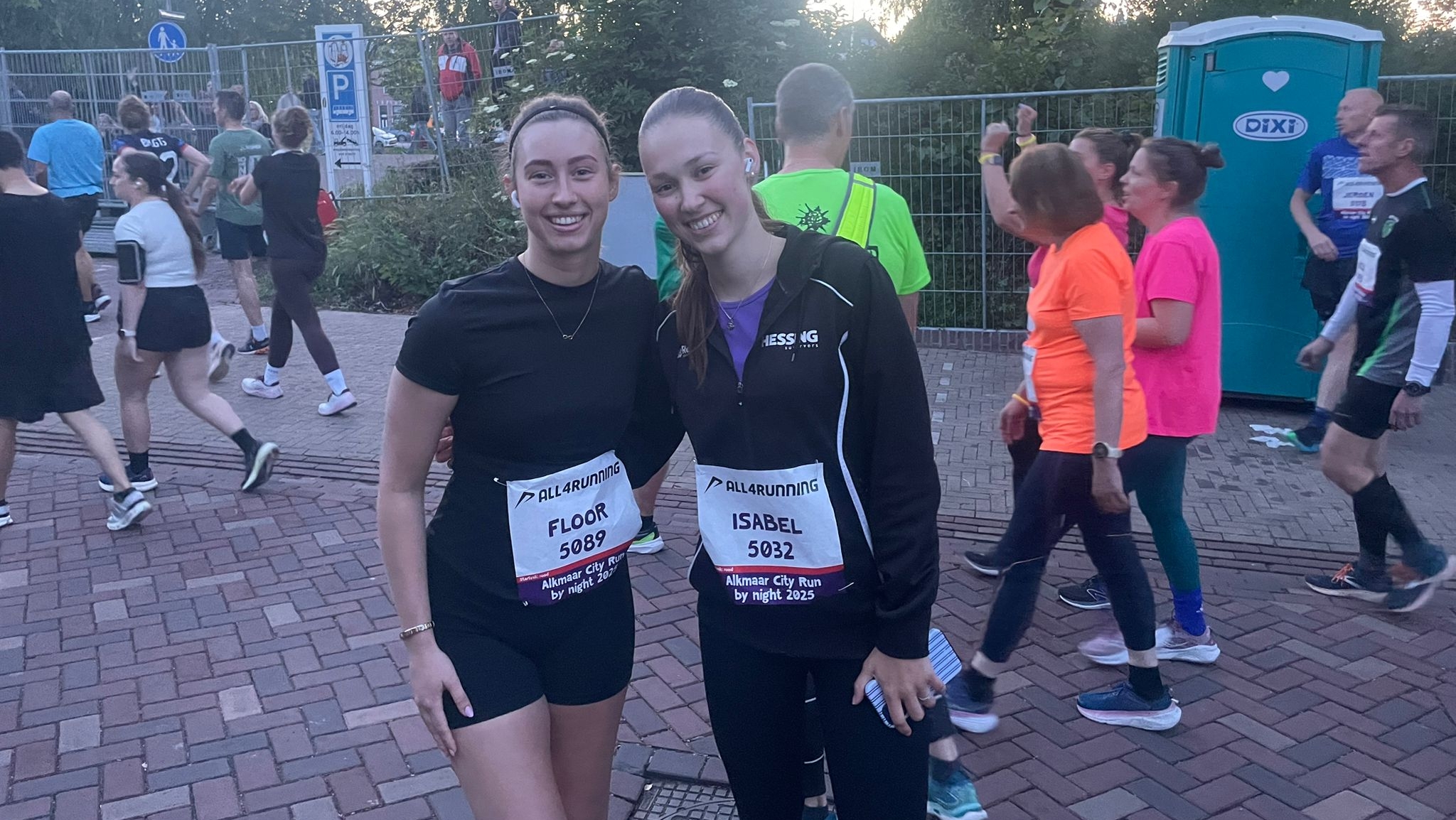 Twee vrouwen glimlachen naar de camera tijdens een hardloopevenement, omgeven door andere deelnemers. Ze dragen sportkleding en startnummers met de tekst 'Alkmaar City Run by night 2023'. Op de achtergrond zijn bomen en een mobiel toilet te zien.