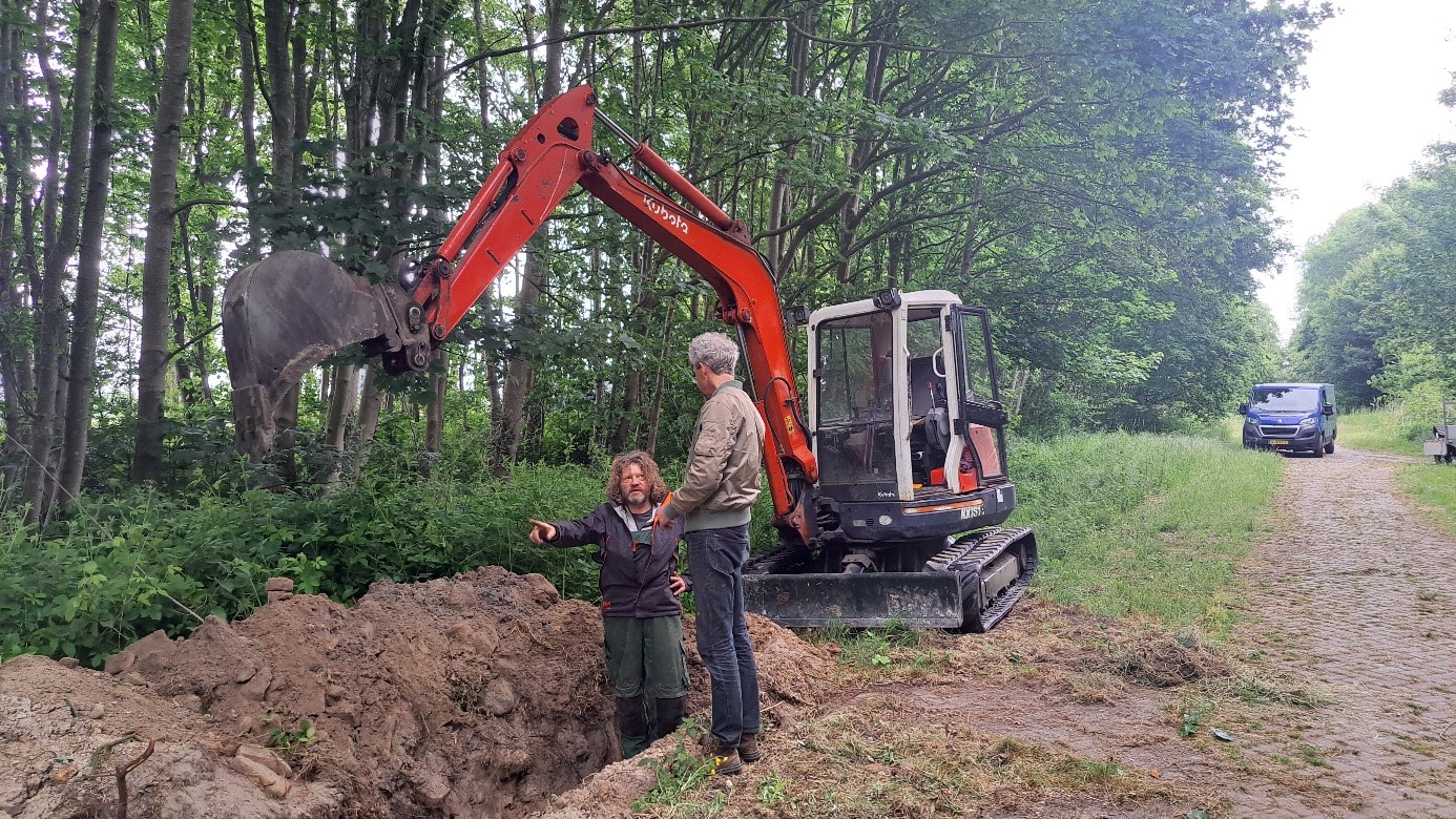Twee personen staan bij een oranje graafmachine in een bosrijke omgeving; de graafmachine is bezig met het uitgraven van een gat naast een onverharde weg waar een bestelwagen geparkeerd staat.