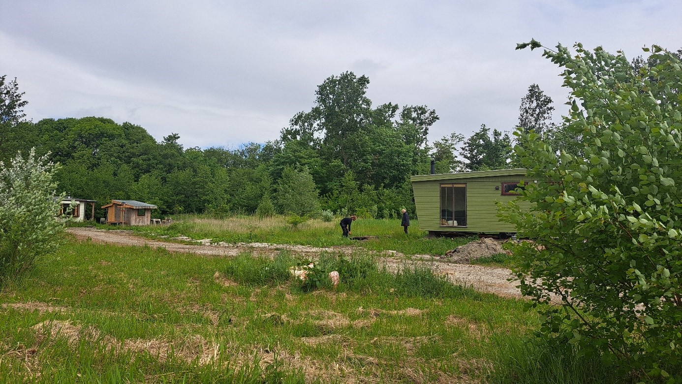 Een groene houten hut op een open veld met enkele mensen bezig in de nabijheid, omgeven door bomen en groene struiken onder een bewolkte hemel.